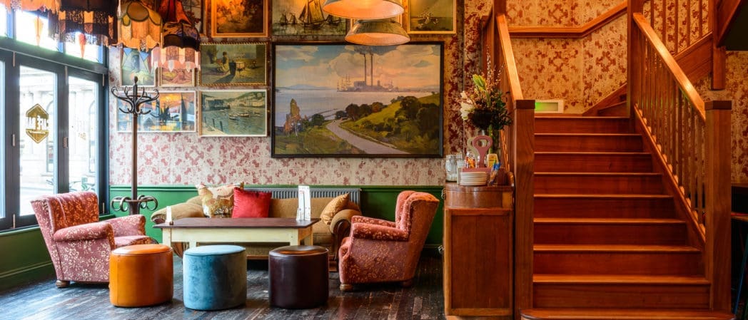 A cozy vintage-style lounge with patterned wallpaper, red armchairs, colorful ottomans, a wooden staircase, and curio-filled shelves, all bathed in the warm glow of hanging lamps and surrounded by various paintings.