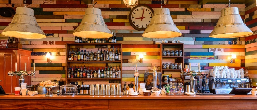 A cozy bar with a colorful wooden plank wall, shelves full of liquor bottles and curios, a coffee machine, pastries on the counter, and three large industrial pendant lights hanging above. A clock hangs on the back wall.
