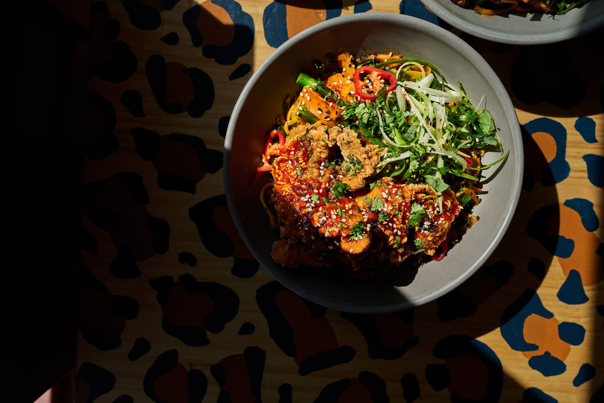 A bowl of noodles topped with crispy fried chicken, fresh herbs, sliced red chilies, and green onions sits on a colorful, animal print-patterned table in bright sunlight.