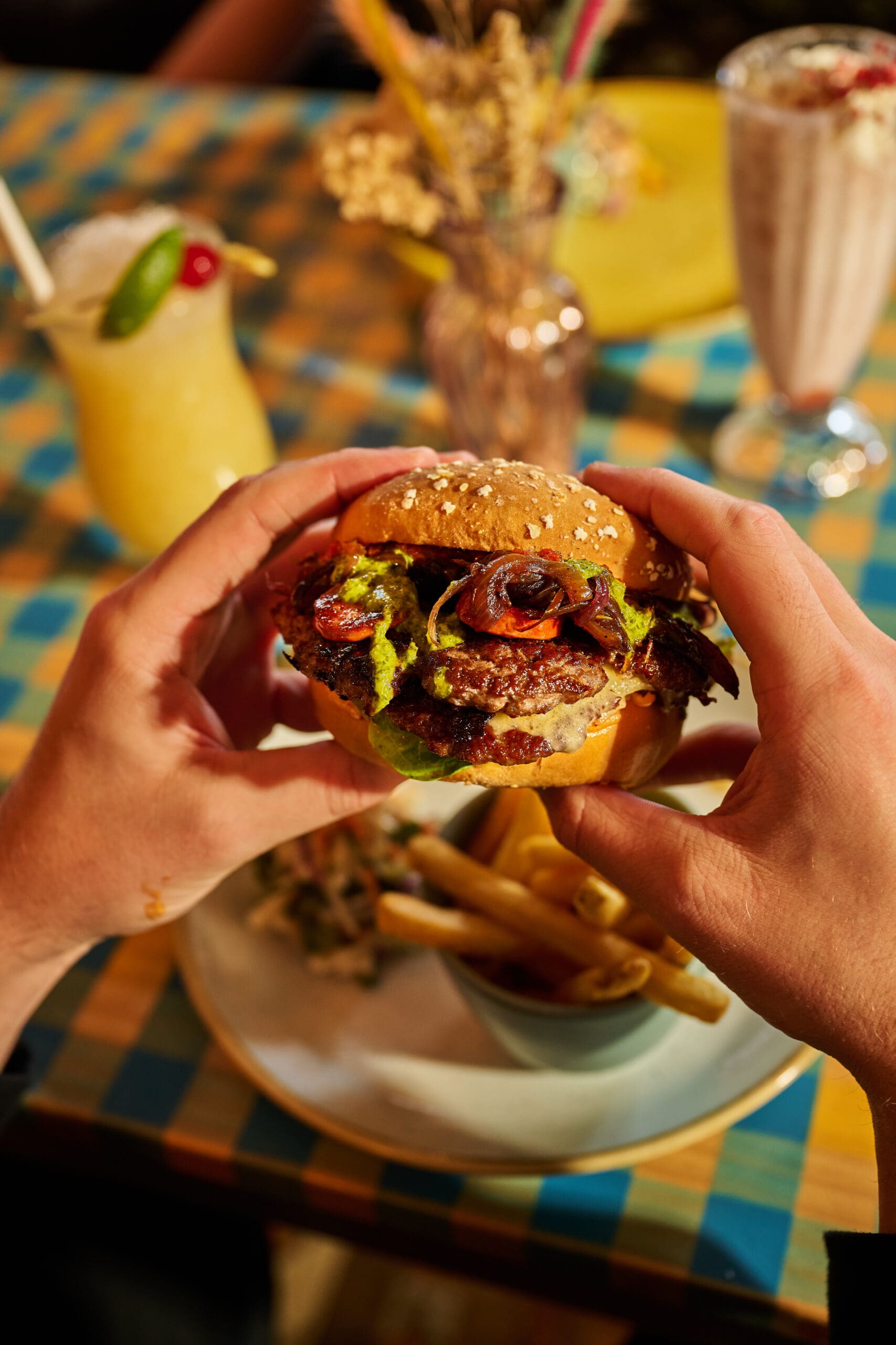 A person holds a gourmet burger with grilled vegetables and leafy greens in both hands above a plate of fries, with drinks and colorful tableware in the background.