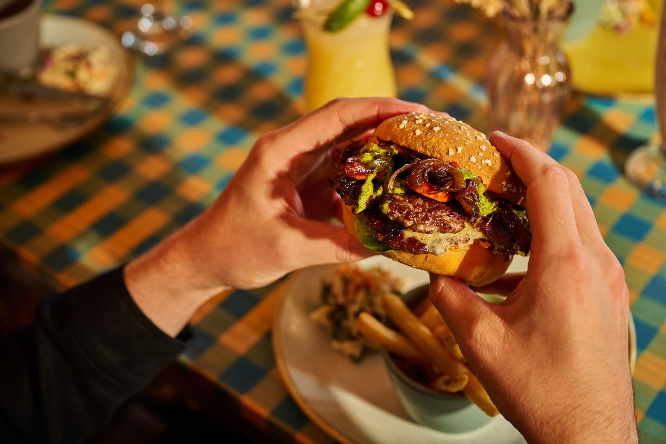 A person holds a sesame seed burger with lettuce, tomato, and sauce above a plate of fries on a table covered with a colorful checkered tablecloth.