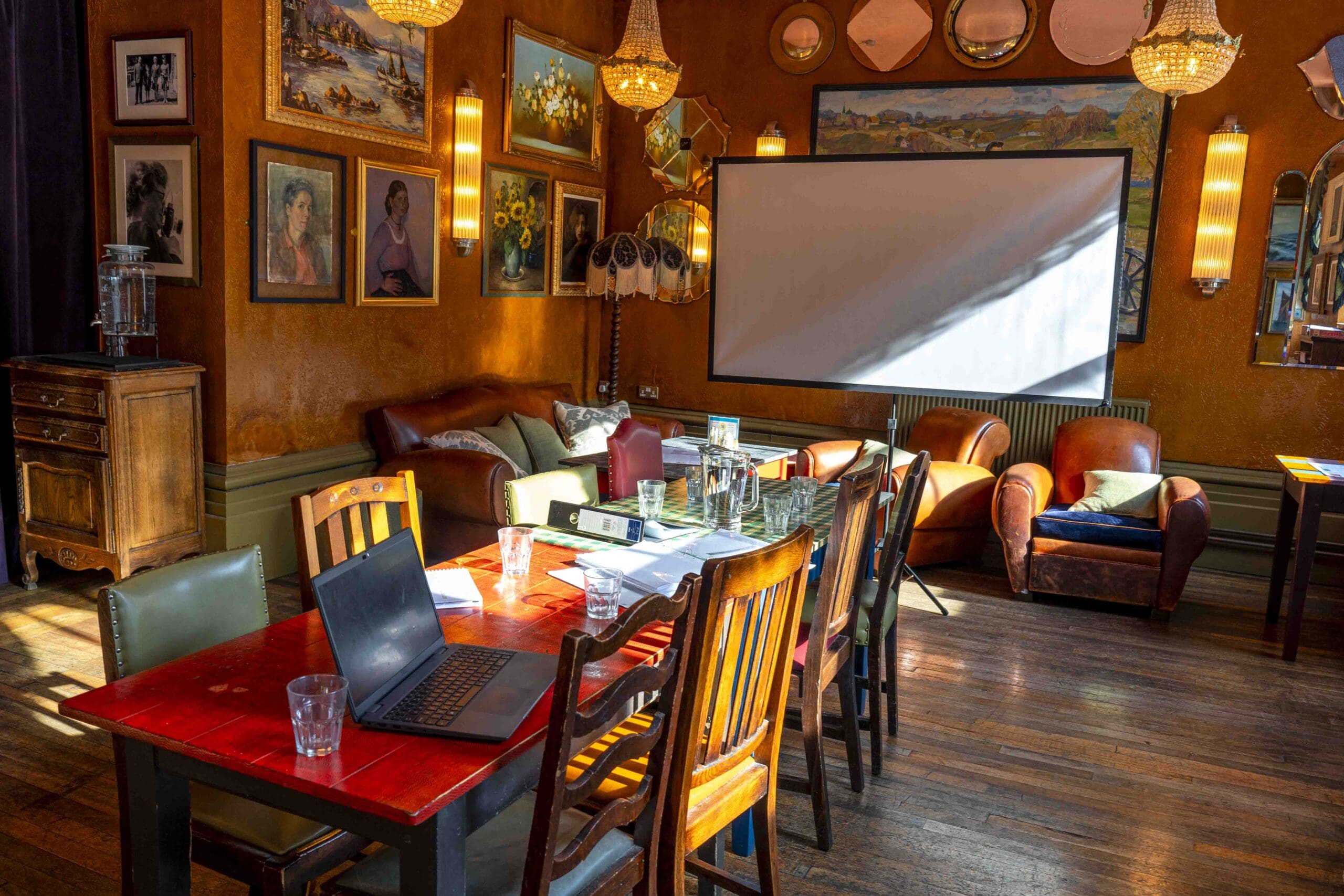 A cozy, warmly lit room with vintage decor, a wooden table with chairs, a laptop, glasses, and papers. There are armchairs, wall art, chandeliers, and a large projector screen with sunlight streaming in.