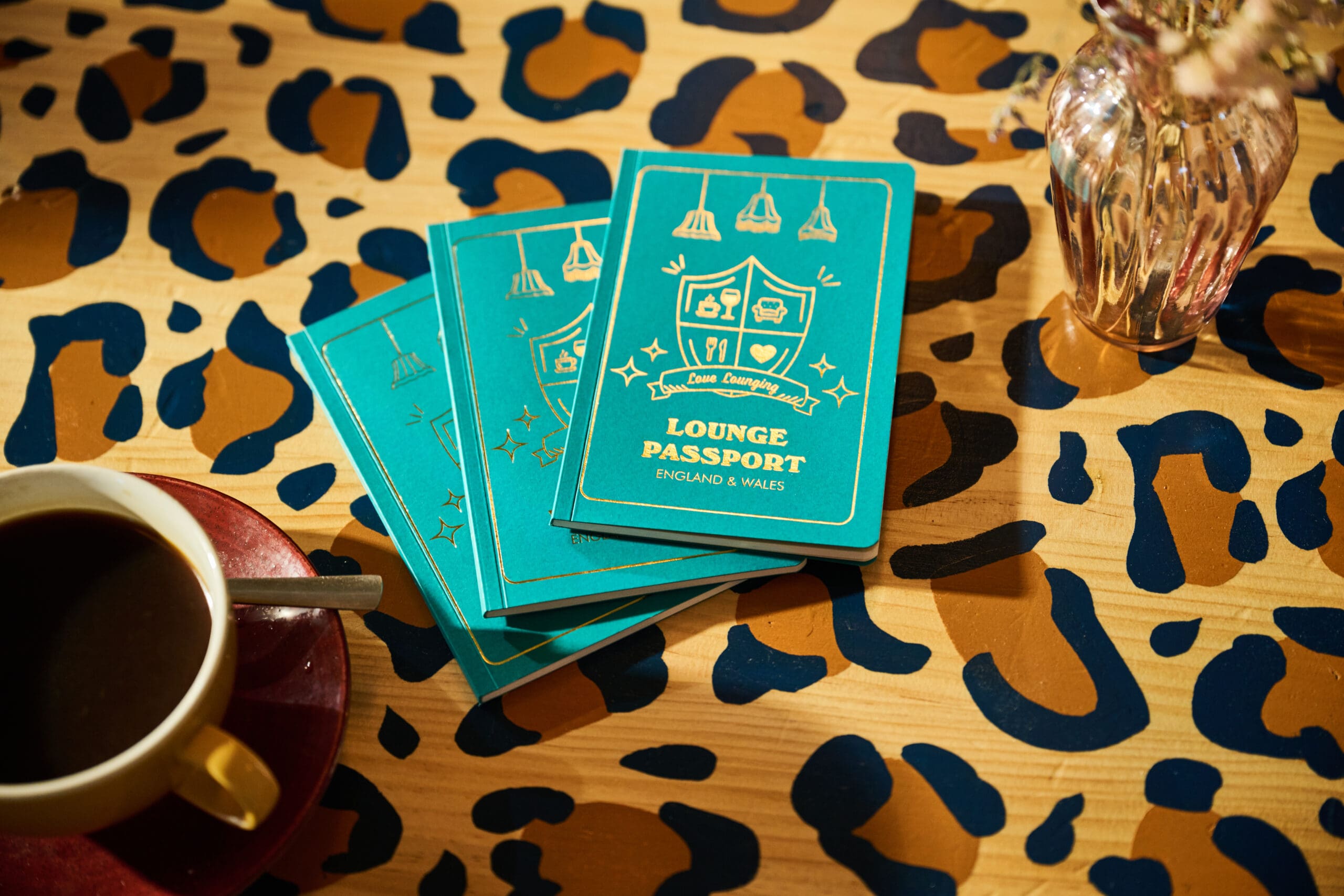 Three teal "Lounge Passport" booklets rest on a wooden table with a bold leopard print pattern, next to a cup of coffee and a small glass vase with dried flowers.