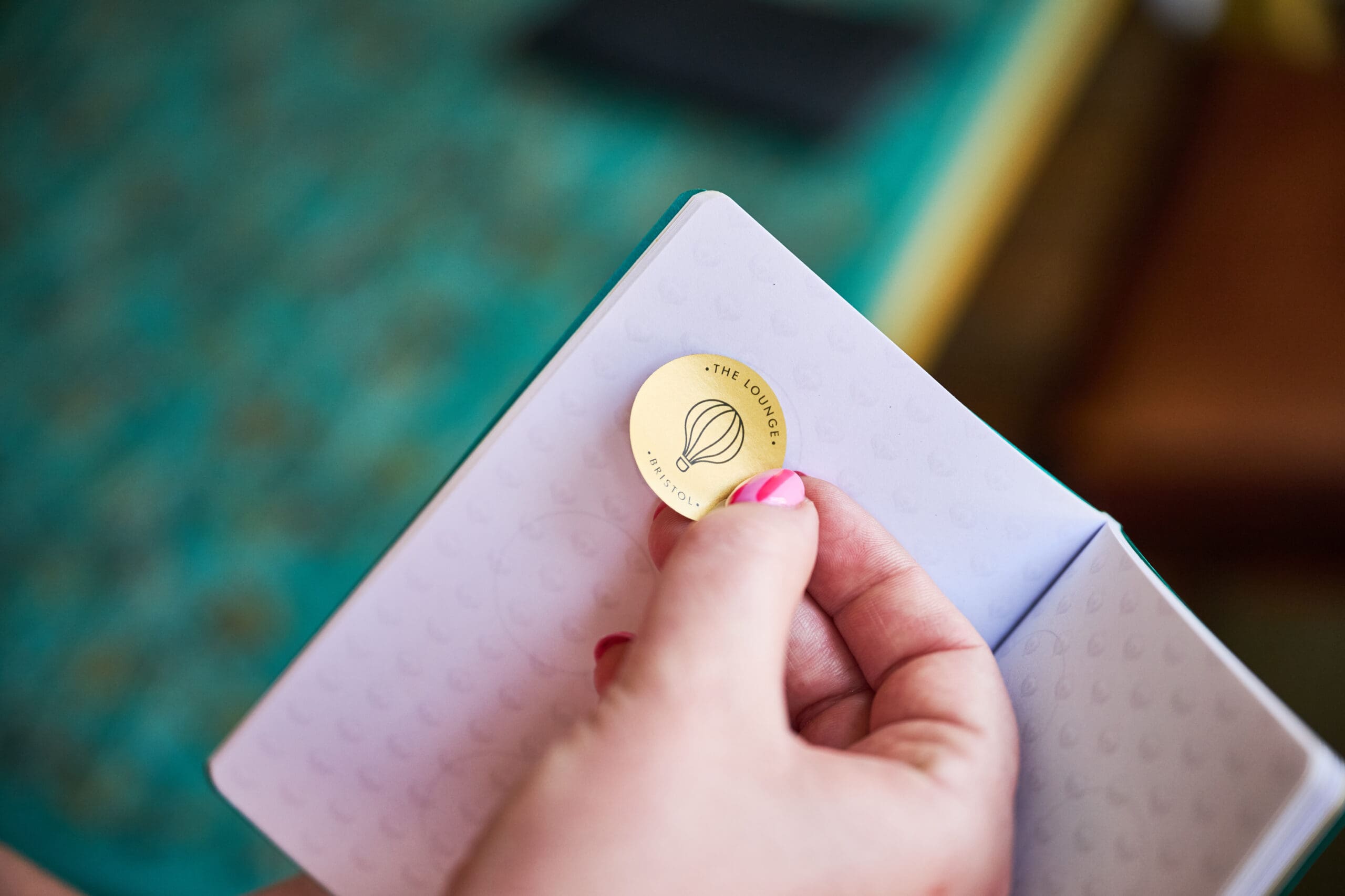 A hand with pink-painted nails places a round gold sticker with a hot air balloon design and the words “THE LOUNGE” on a white notebook with a subtle heart pattern.
