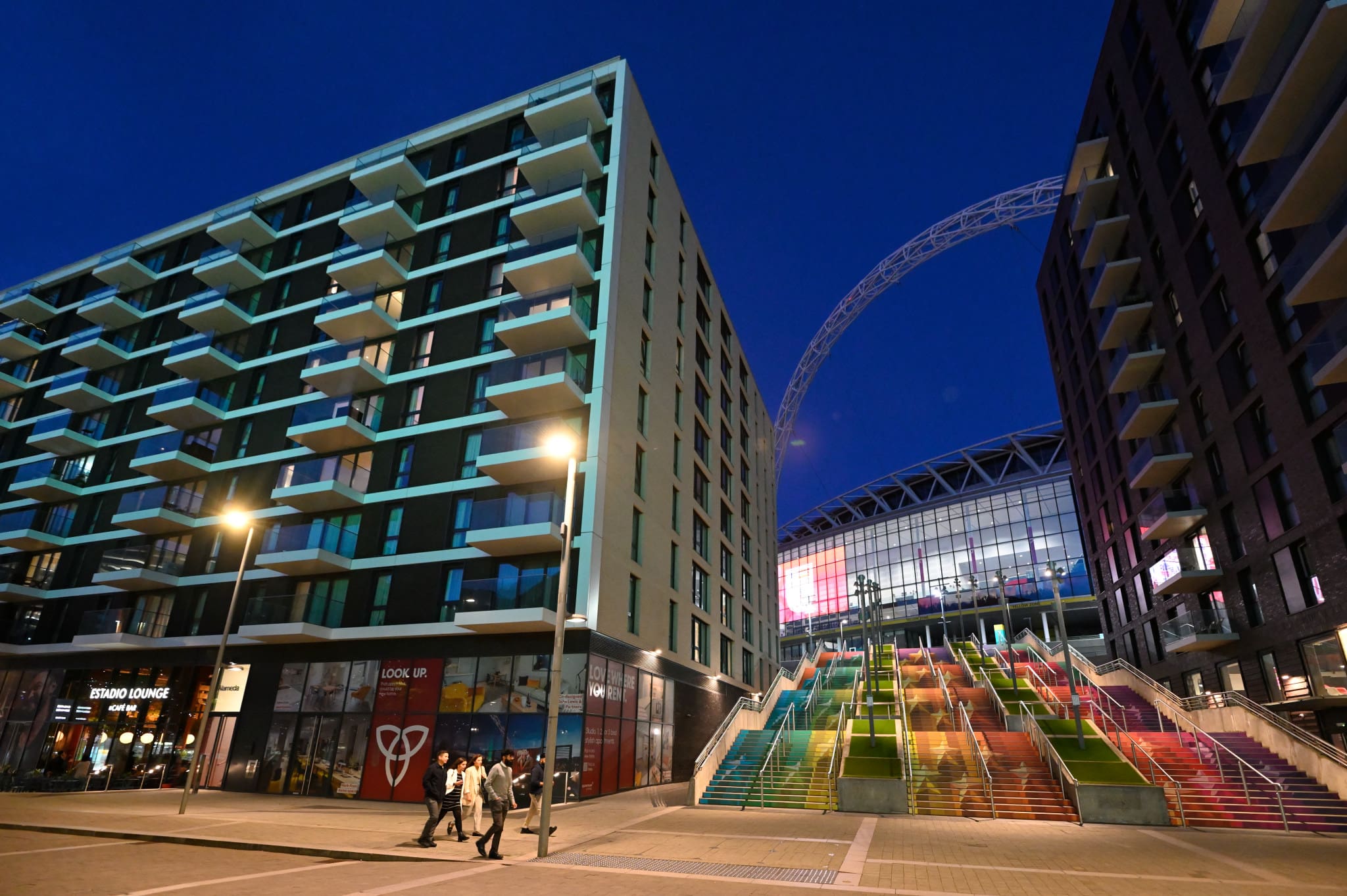 Modern apartment buildings and a group of people walking near illuminated, rainbow-colored stairs leading towards Wembley Stadium, with its iconic arch visible in the night sky.