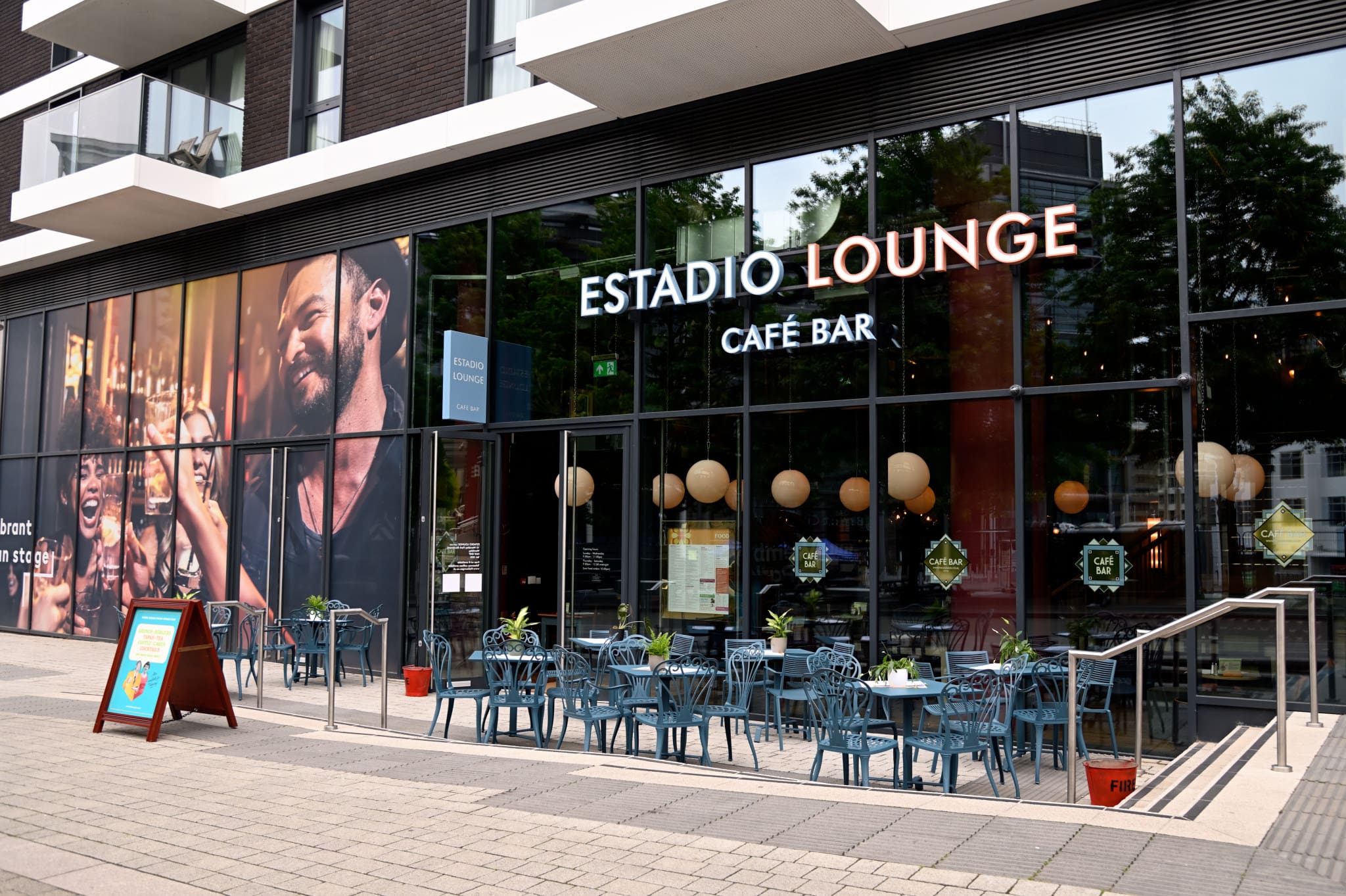 Outdoor seating area with blue tables and chairs in front of Estadio Lounge café bar. Large windows showcase the lively Estadio interior and a mural of people smiling and drinking. Signs and plants decorate the entrance.