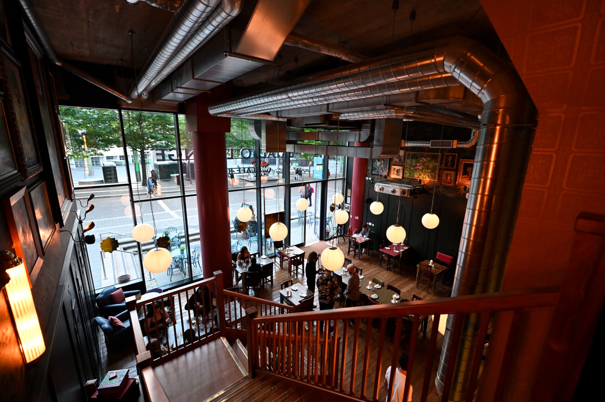 A cozy, modern café near Estadio with large windows, hanging globe lights, exposed ductwork, and wood accents, viewed from a staircase above. People are seated at tables, and trees are visible outside.