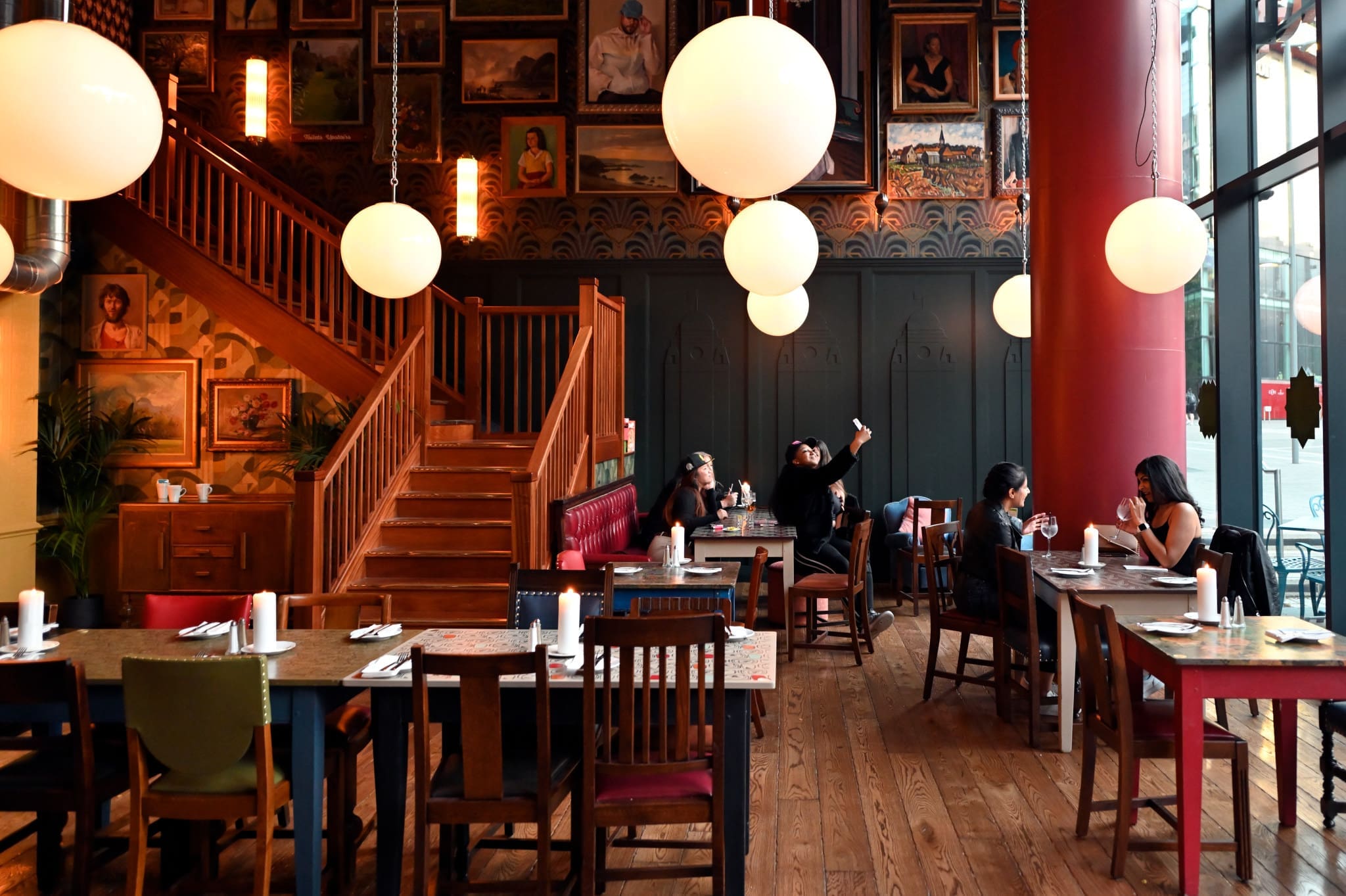 A stylish restaurant near Estadio, with wooden floors, eclectic chairs, large round lights, and framed pictures on the wall. Four people sit at a table, one raising their phone—perhaps snapping a photo of the moment.