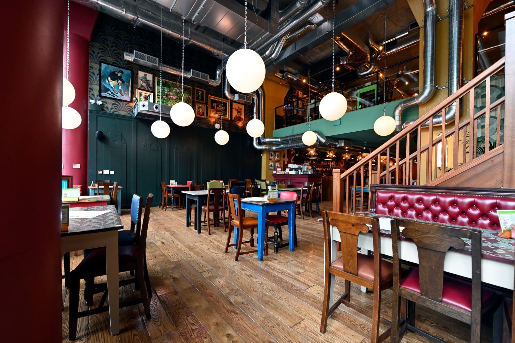 A brightly lit, modern restaurant near Estadio with wooden floors, colorful chairs, round hanging lights, eclectic wall art, and an open staircase. Tables are set for dining in an inviting and stylish atmosphere.