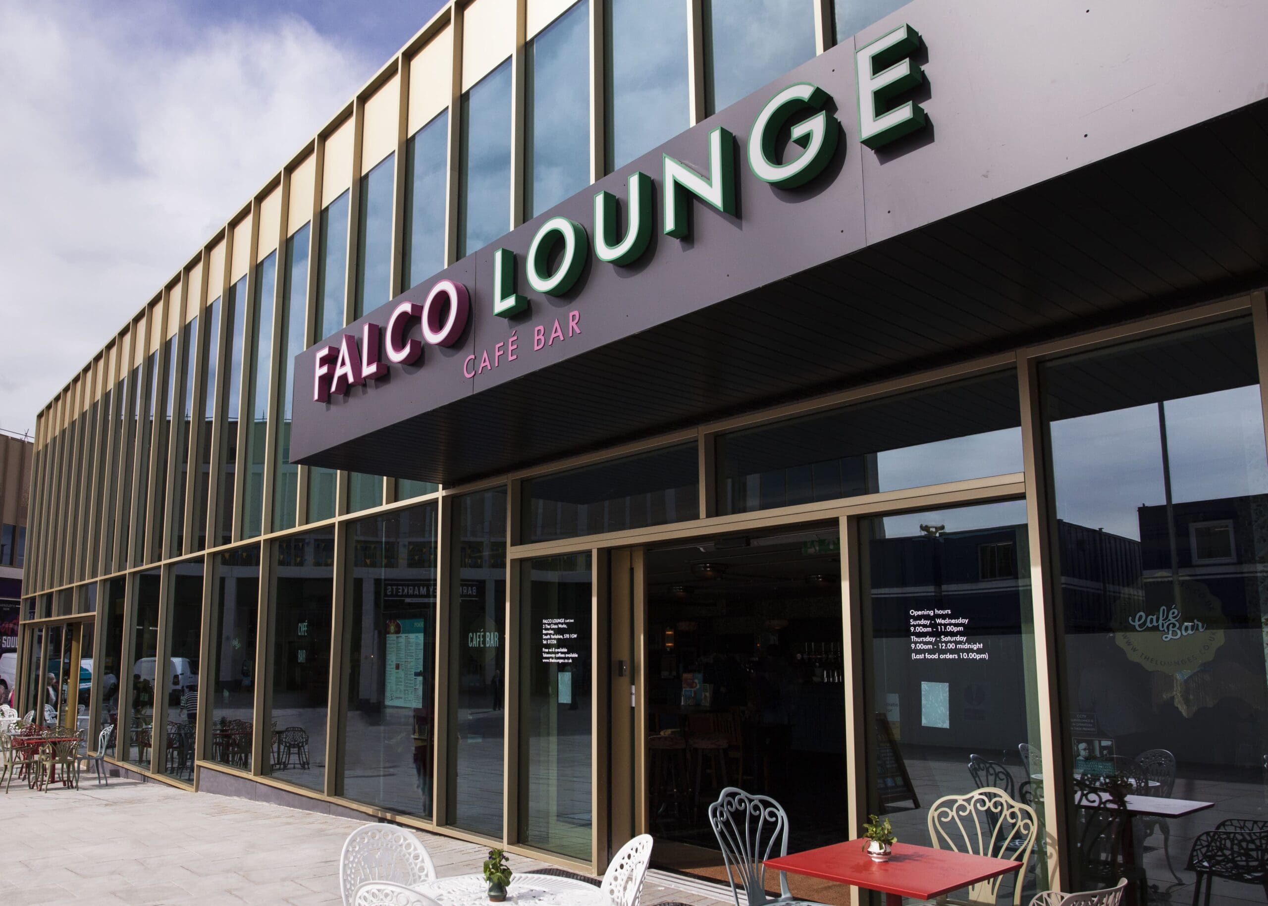 Exterior view of Falco Lounge café bar with large glass windows, outdoor seating featuring white and red chairs and tables, and a modern building design under a partly cloudy sky invites you to experience Falco’s relaxing atmosphere.