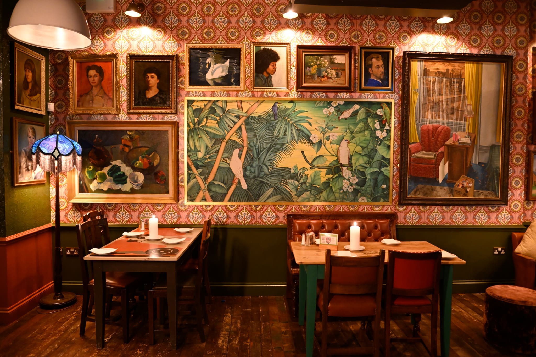 A cozy restaurant interior with vintage wallpaper, wooden tables and chairs, and walls adorned with eclectic framed paintings, including portraits and a large tropical bird mural. Warm ferro lighting creates a welcoming atmosphere.