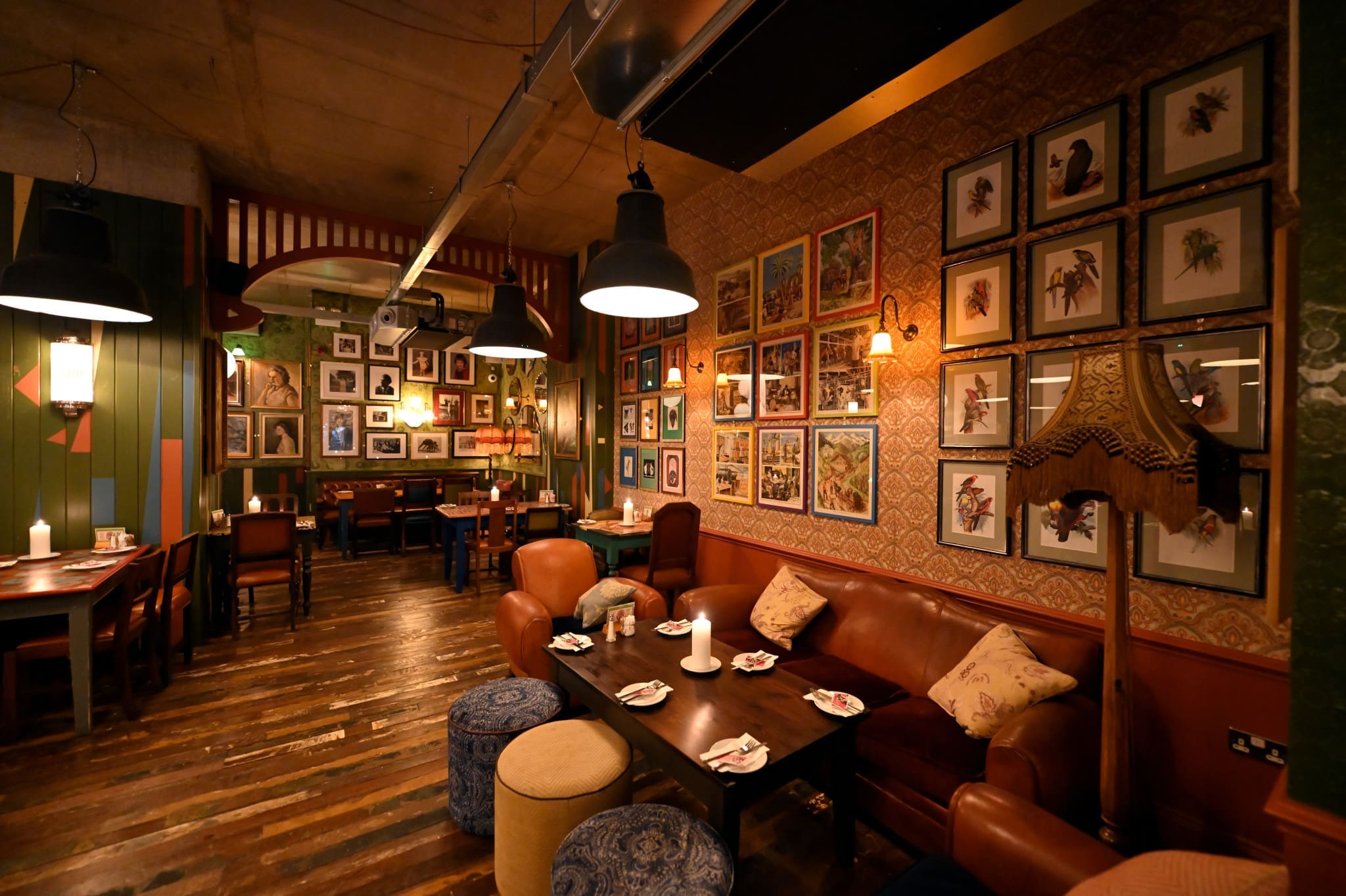 Cozy restaurant interior with warm lighting, leather couches, Ferro accents, wooden tables, patterned wallpaper, vintage lamps, and walls covered with framed art and photographs. Wooden floor and eclectic decor create an inviting, intimate atmosphere.