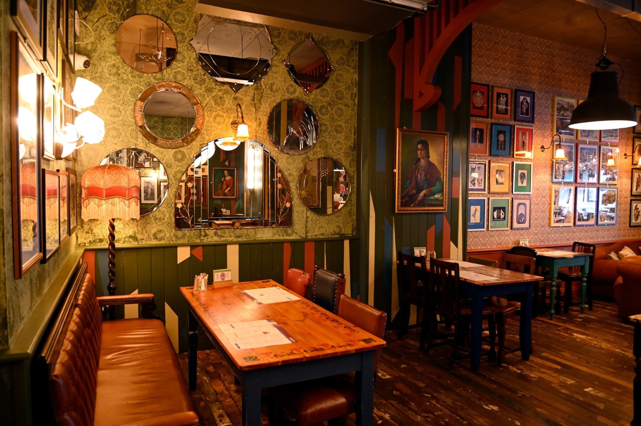 A cozy, eclectic restaurant interior with vintage decor, leather seating, wooden tables, Ferro accents, patterned wallpaper, round mirrors, framed photos, and warm ambient lighting.