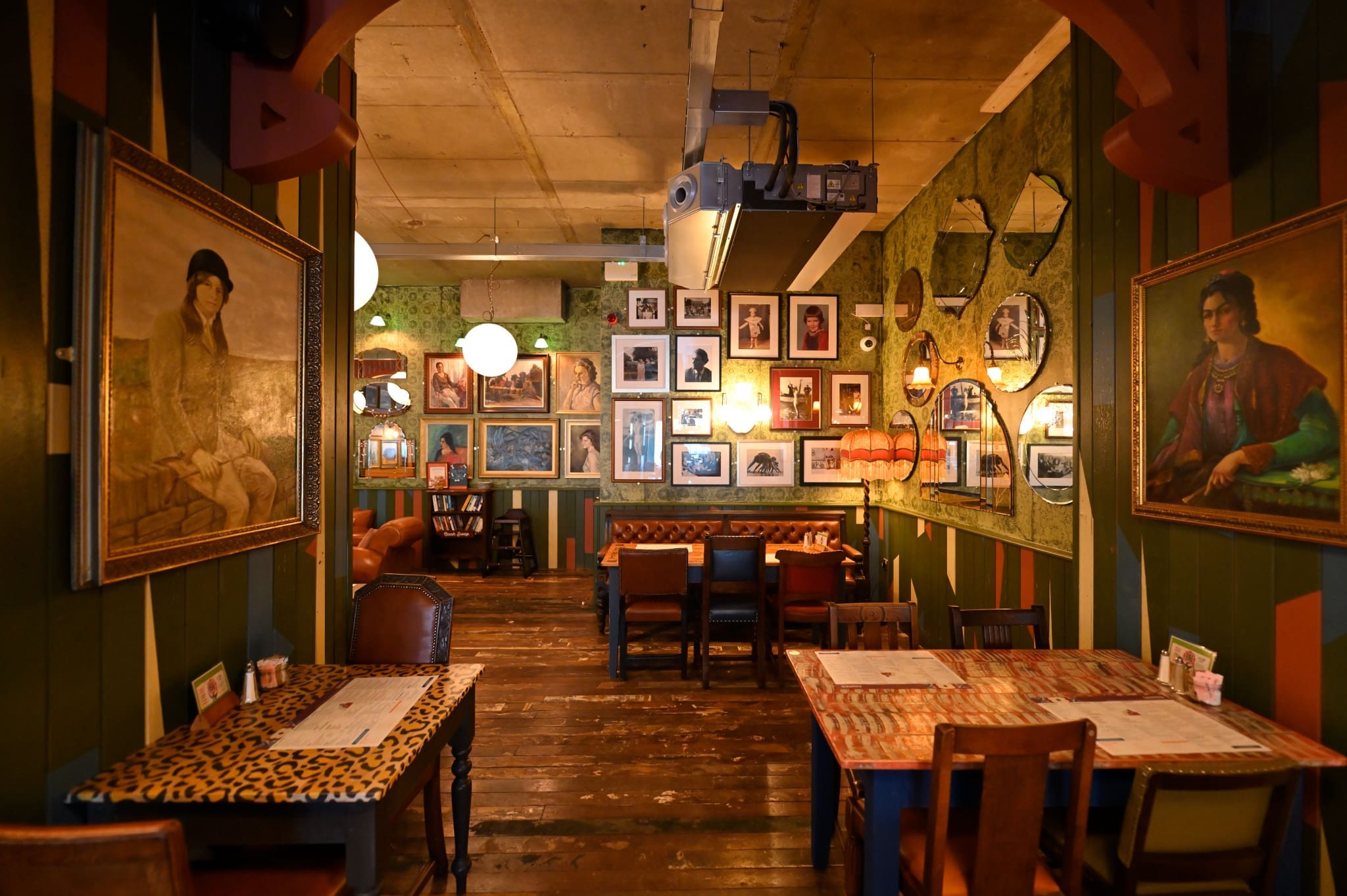 A cozy, eclectic restaurant interior with wooden floors, colorful Ferro tables and chairs, and walls adorned with various framed paintings, photographs, and mirrors. Warm lighting creates an inviting atmosphere.