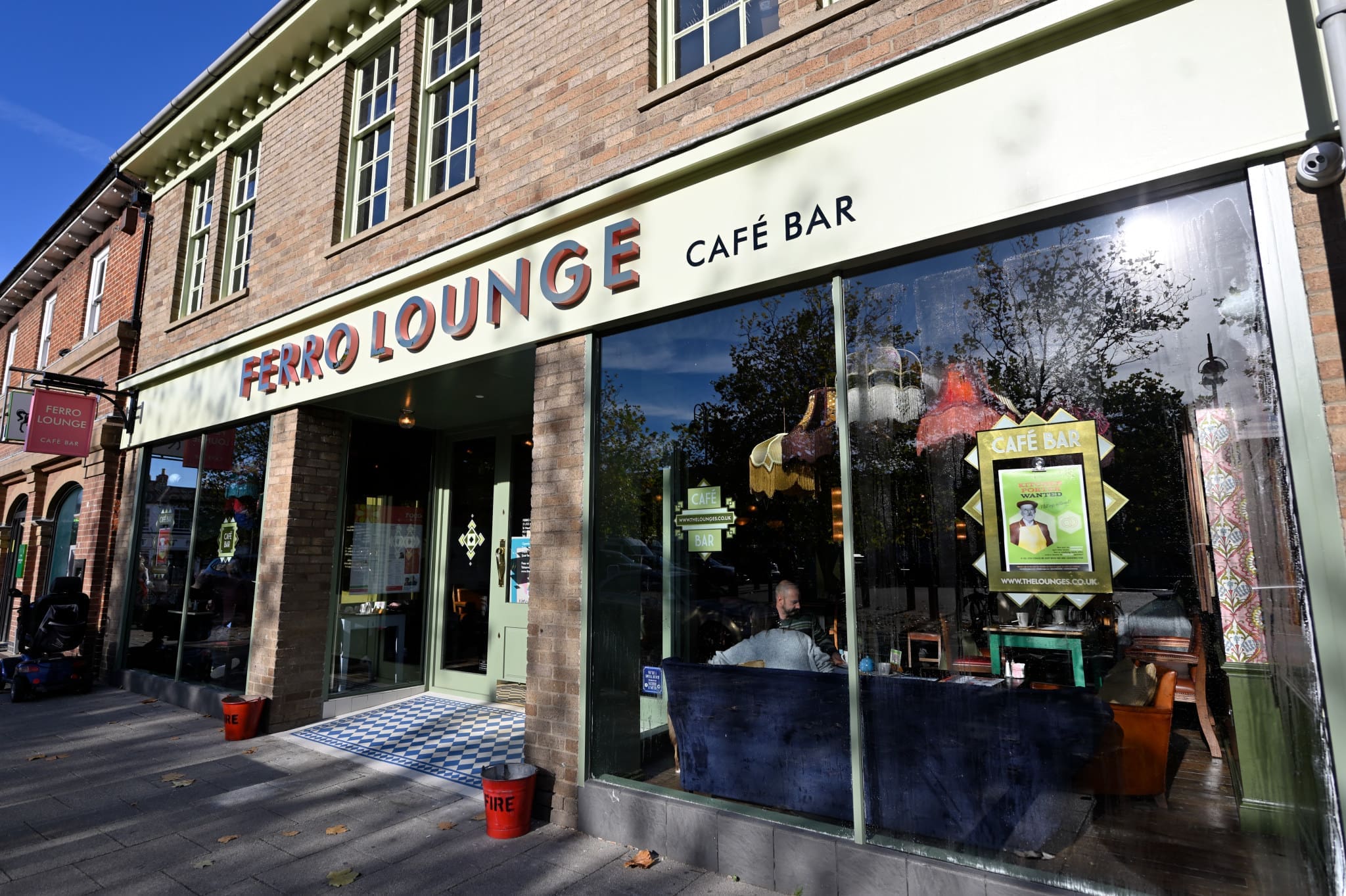 Street view of Ferro Lounge Café Bar with large windows showing people enjoying the inviting Ferro atmosphere inside. The brick building features “Ferro Lounge” signage above the entrance and reflections of trees in the glass.