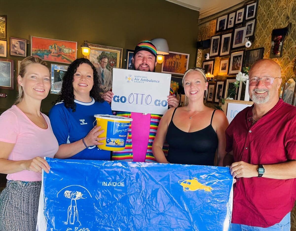 Five smiling people pose indoors, holding Air Ambulance-themed signs and a blue banner. One person wears a rainbow shirt and hat. The background features framed pictures on the wall.