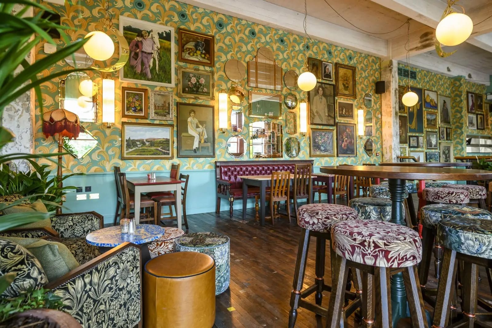 Eclectic Torino cafe interior with patterned wallpaper, vintage framed artwork, round hanging lights, cushioned seating, stools with ornate upholstery, wooden tables, and plants creating a cozy, artistic atmosphere.