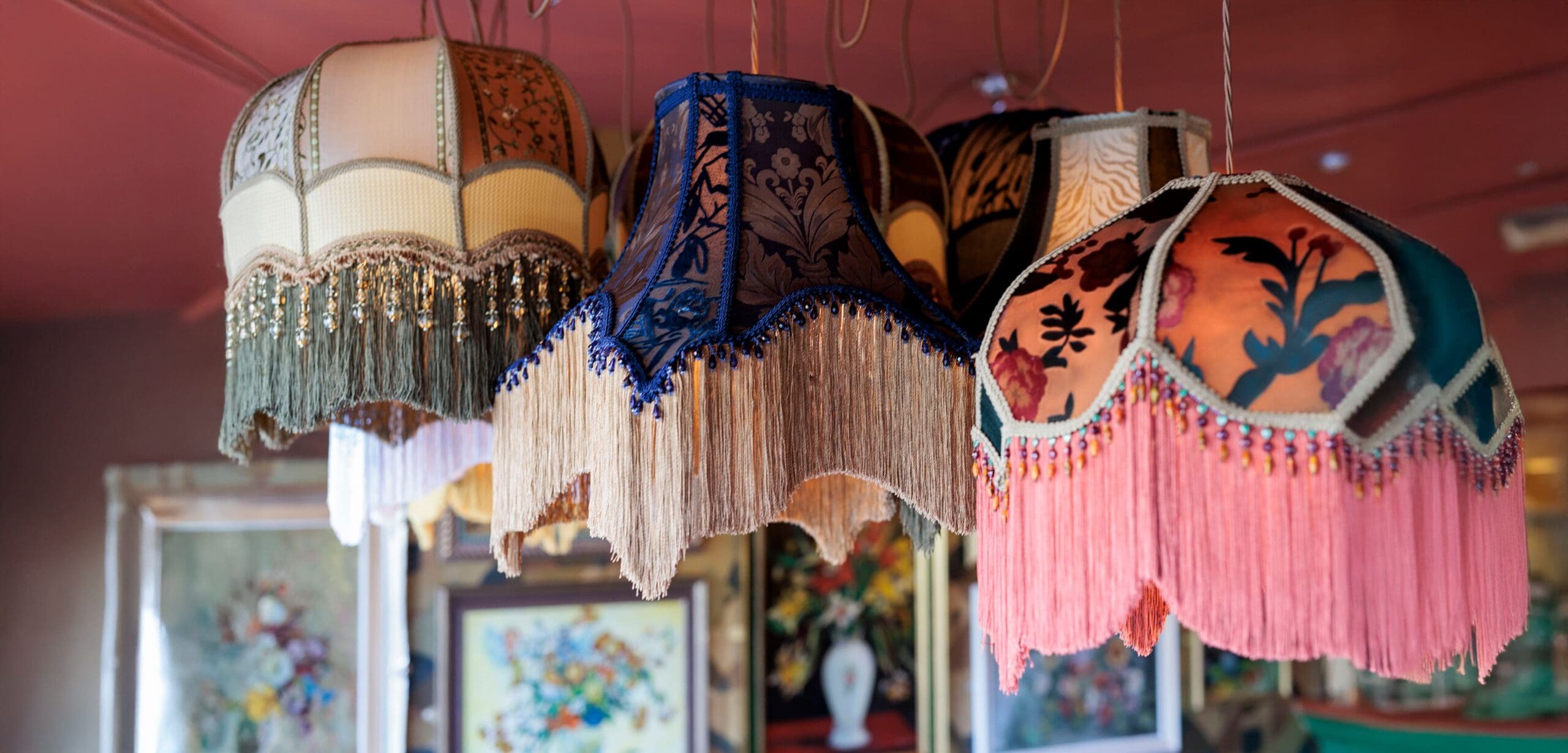 A group of vintage fabric lampshades with colorful fringe hangs from the ceiling in our About Us space, set against a room adorned with framed floral artwork in the background.