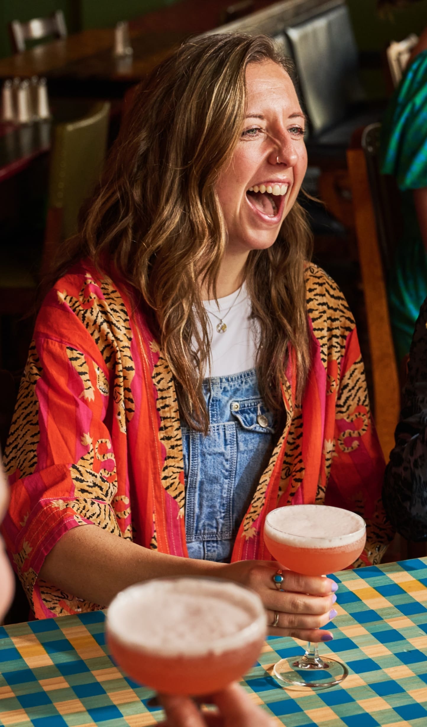 A woman with wavy hair, wearing a red patterned jacket and denim overalls, laughs while holding a pink cocktail at a checkered table in lively Torino.