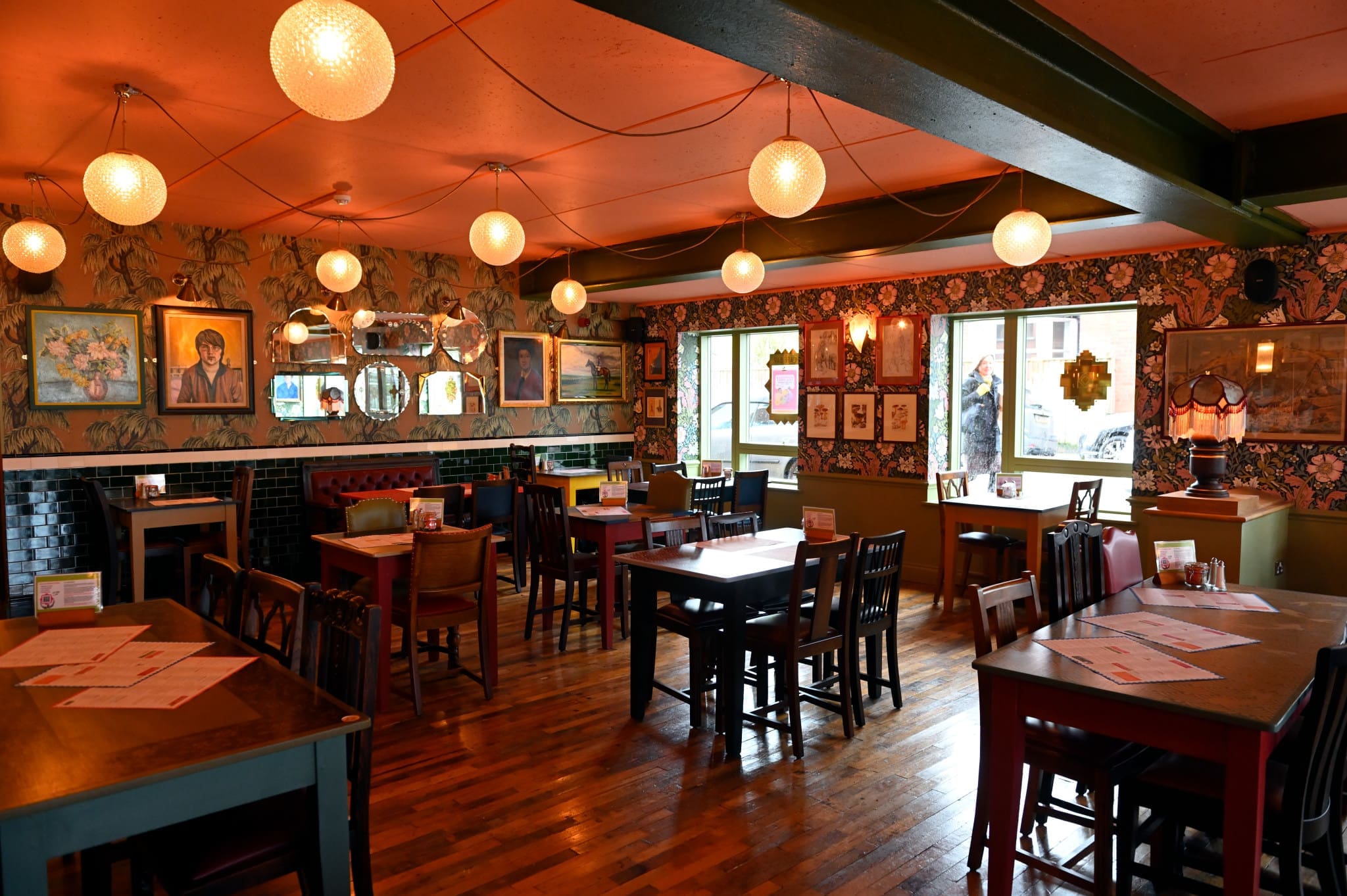 Fioro is a cozy, vintage-style restaurant with wooden floors, mismatched tables and chairs, globe pendant lights, floral wallpaper, framed art on the walls, and natural light streaming through the windows.