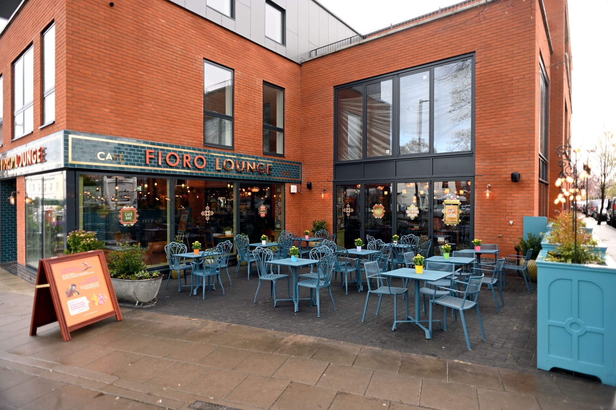 Fioro Lounge's outdoor seating area features blue tables and chairs on a patio before a modern red-brick building, with potted plants and a sidewalk sandwich board adding charm to the Fioro experience.