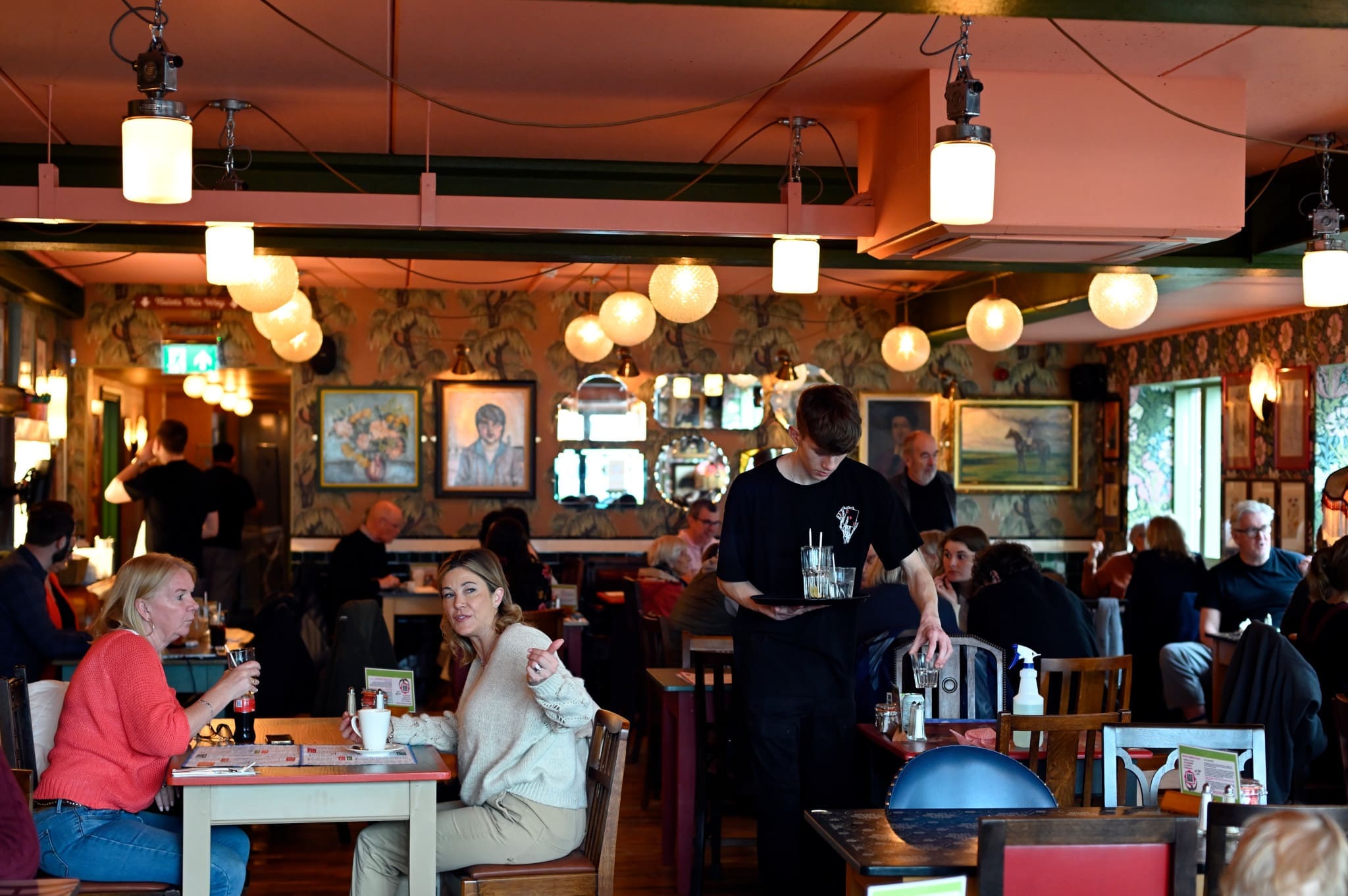 A busy Fioro restaurant interior with people eating and talking at tables. A waiter carries drinks, and warm hanging lights and paintings decorate the cozy, colorful space.