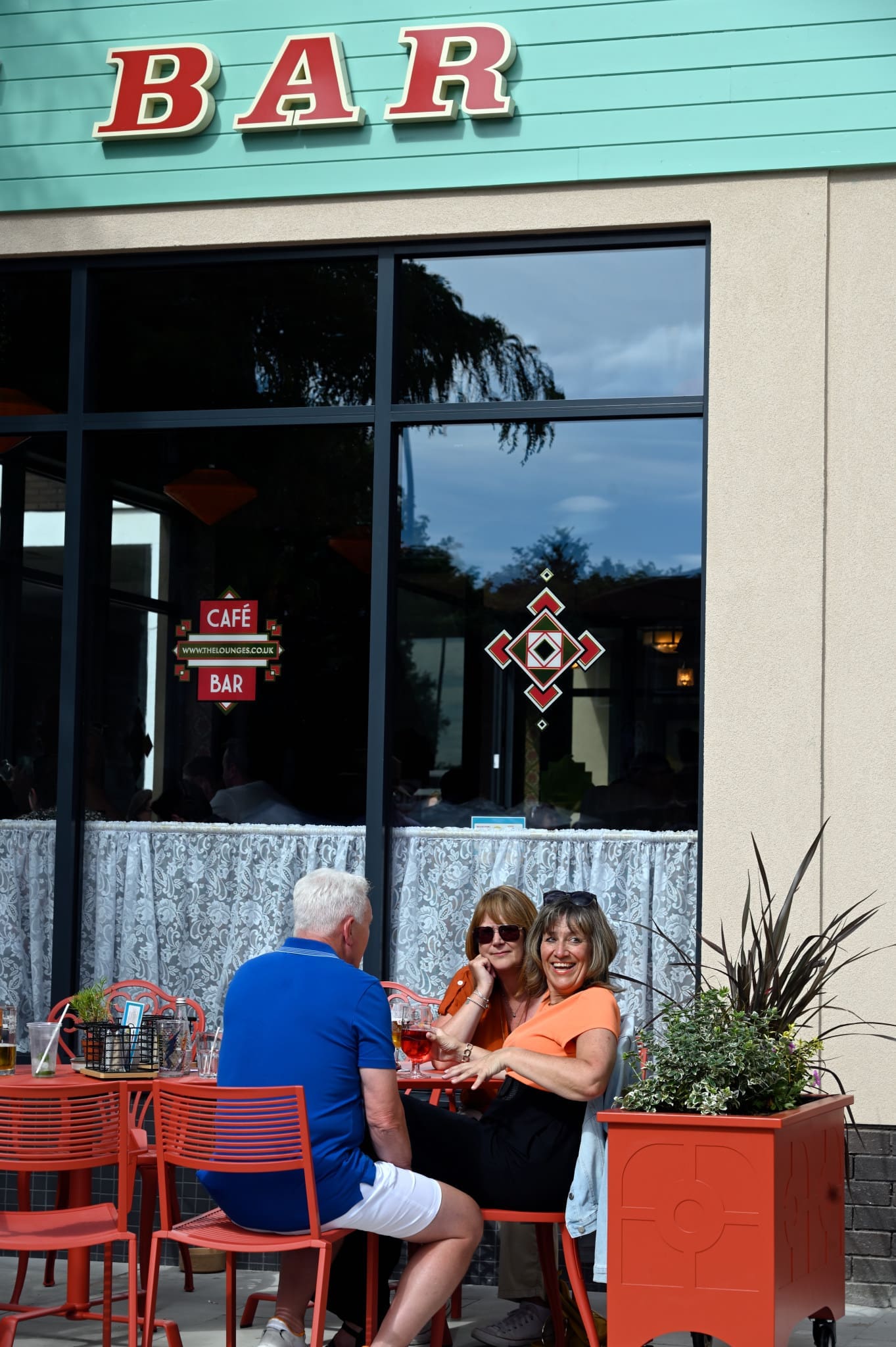 Three people sit at a red outdoor table outside Fossato, smiling and enjoying drinks. The bar sign is visible above large windows, with a large red planter filled with green foliage nearby.
