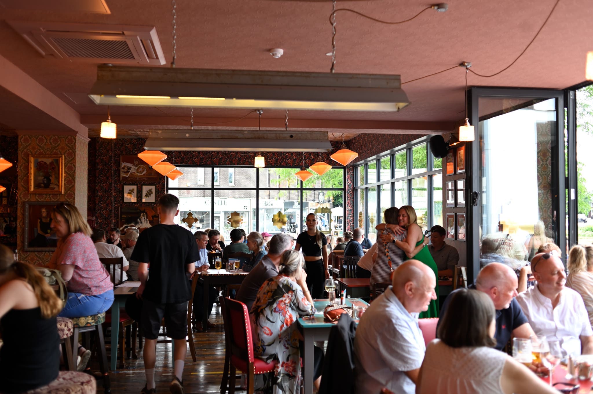 A busy, lively restaurant with people sitting at tables, eating, drinking, and talking. Large windows let in natural light, while warm fossato-style lamps hang from the ceiling. Some people stand near the entrance; others are seated.