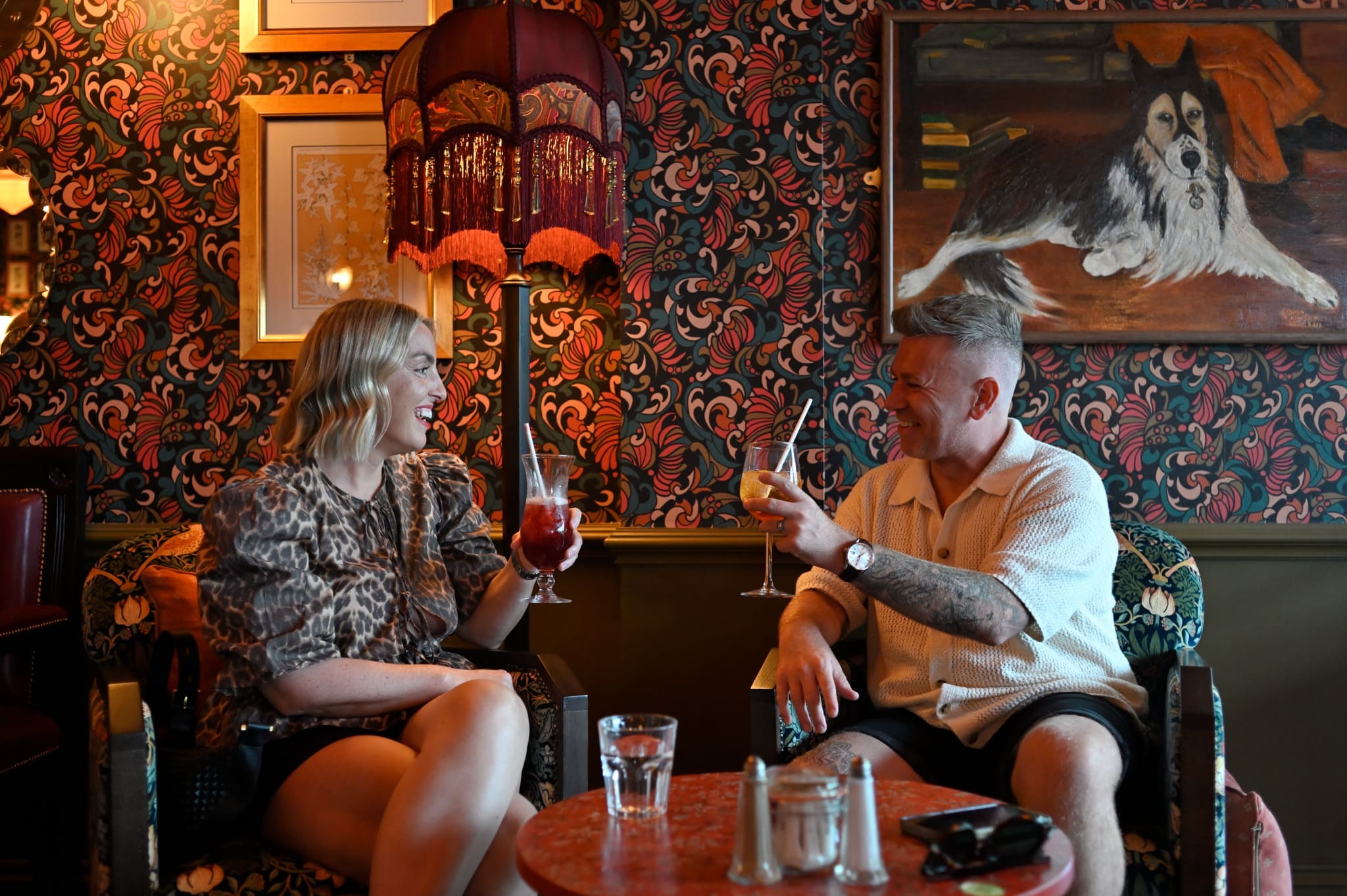 Two people sit in a cozy, vintage-style room with floral wallpaper, clinking cocktail glasses and smiling. A painting of a dog hangs on the wall above a fossato-inspired lamp with a fringed shade, casting warm light over the inviting scene.
