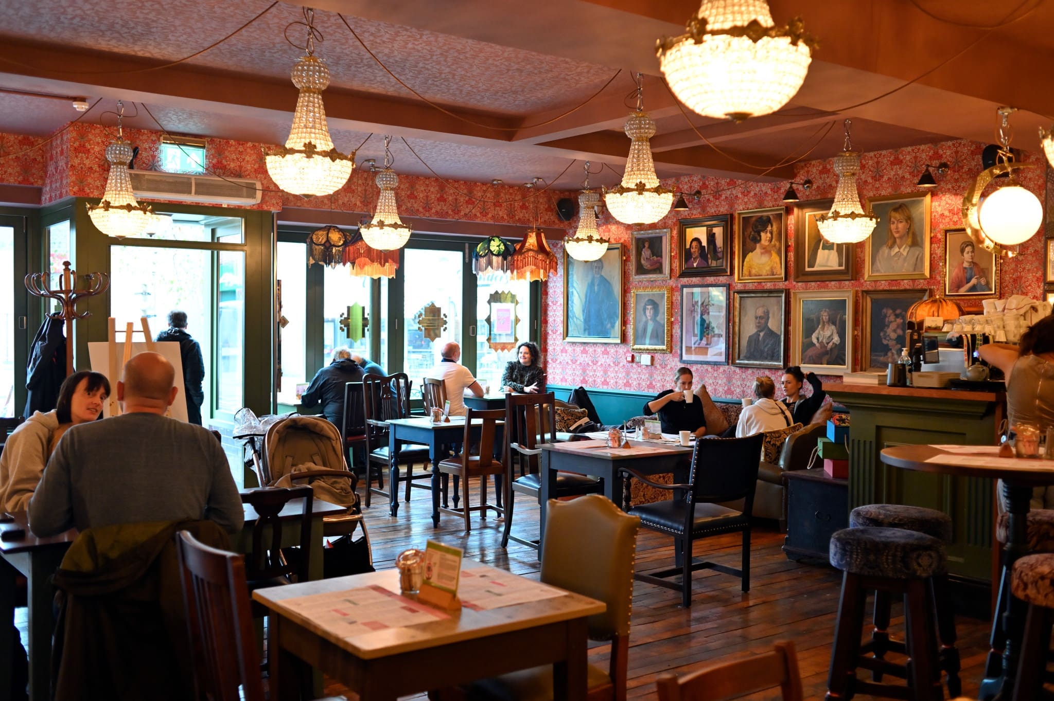 A cozy café in Francisco with chandeliers, floral wallpaper, and framed portraits on the walls. Several people sit at tables, chatting and relaxing. The atmosphere is warm and inviting with natural light streaming through large windows.