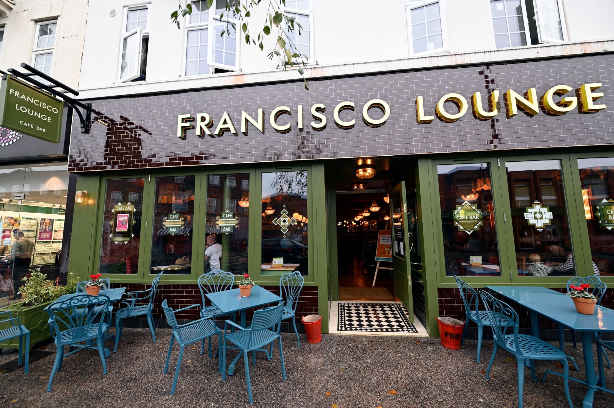 The exterior of Francisco Lounge café bar features green-framed windows, outdoor teal tables and chairs, potted plants, and open doors warmly inviting customers inside Francisco for a relaxing visit.