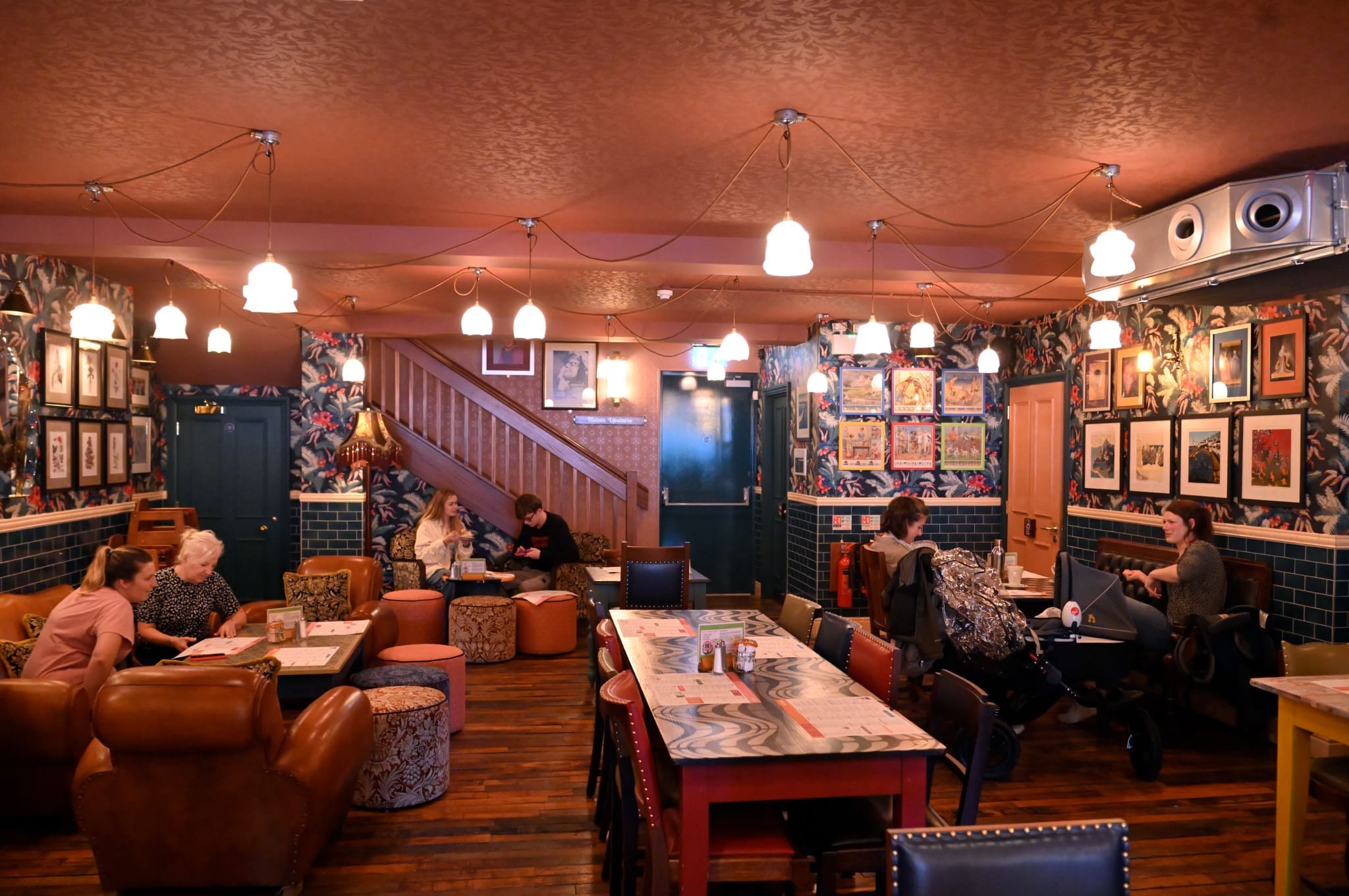 A cozy, eclectic café in Francisco with warm lighting, patterned wallpaper, framed art, and a mix of armchairs and communal tables. Several people are seated, talking, reading, or working, creating a relaxed, inviting atmosphere.