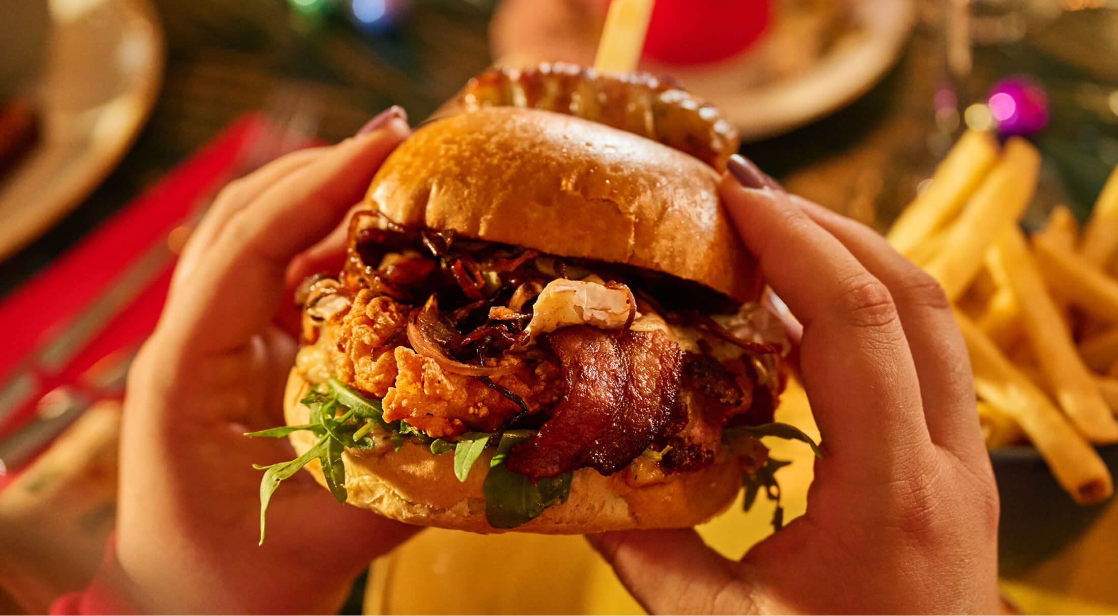 A person holds a large burger filled with fried chicken, bacon, lettuce, and onions in a shiny bun. A plate of French fries is in the background, suggesting a meal setting.
