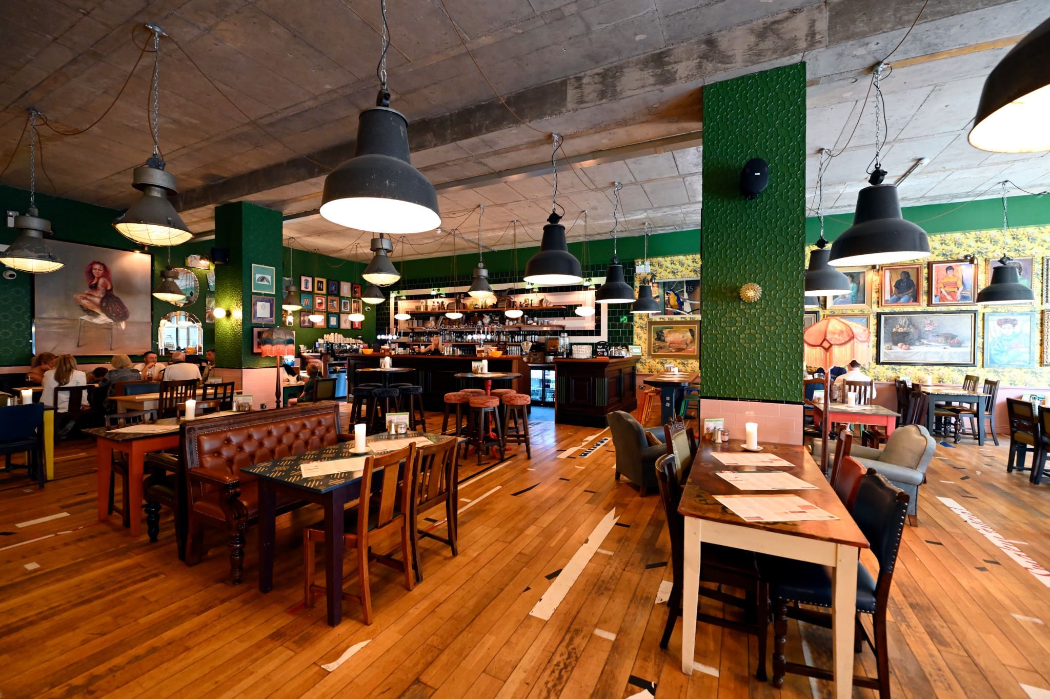 A cozy, modern restaurant with wooden floors, eclectic furniture, and hanging industrial lights sets the mood at Gallico. Walls decorated with framed artworks and green accents complete the ambiance as guests enjoy their meals in the background.