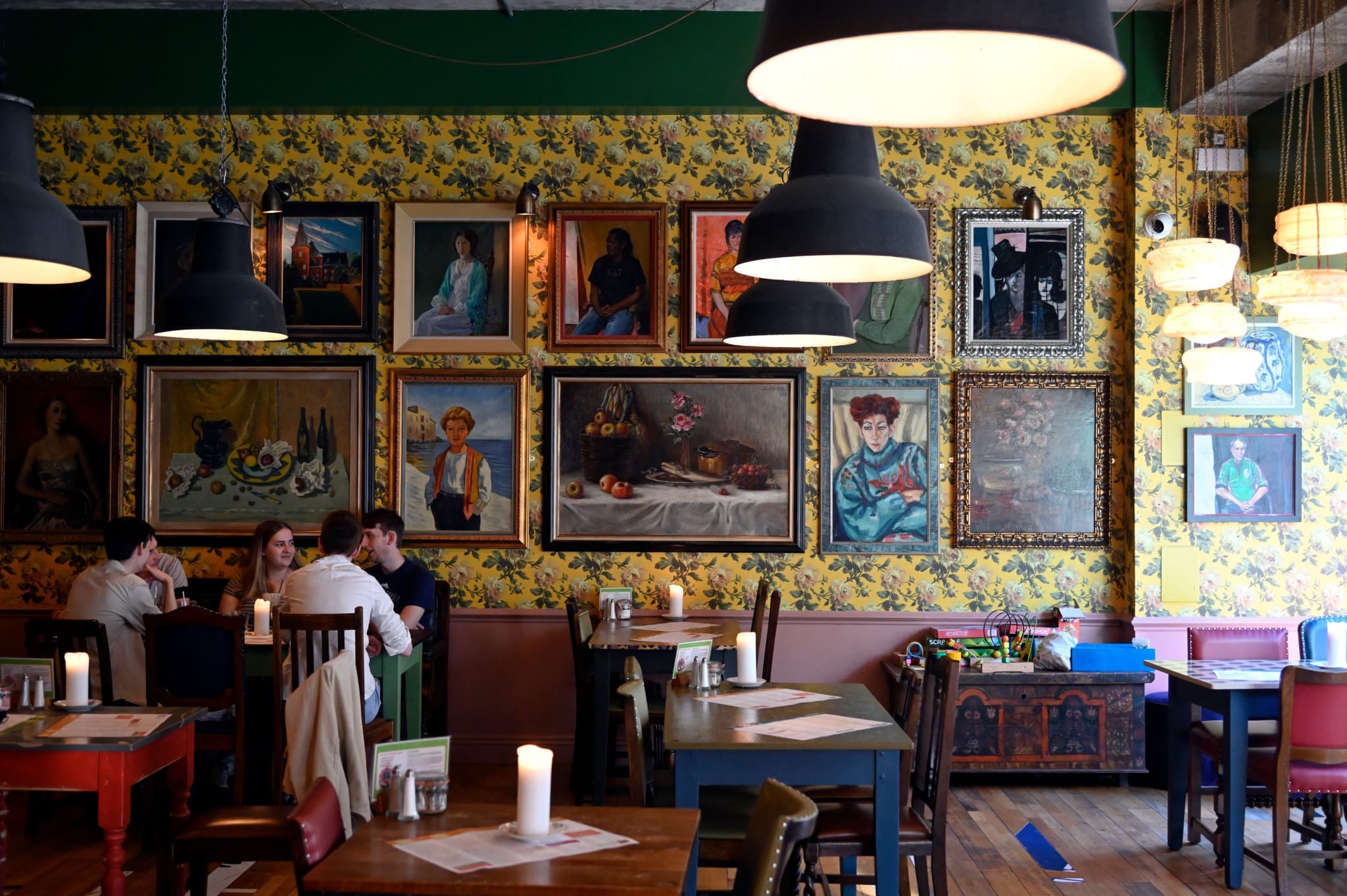 A cozy restaurant with eclectic vintage decor and Gallico charm, featuring floral wallpaper and framed paintings. Groups gather at wooden tables under hanging lamps, as candles add warm light to the inviting scene.