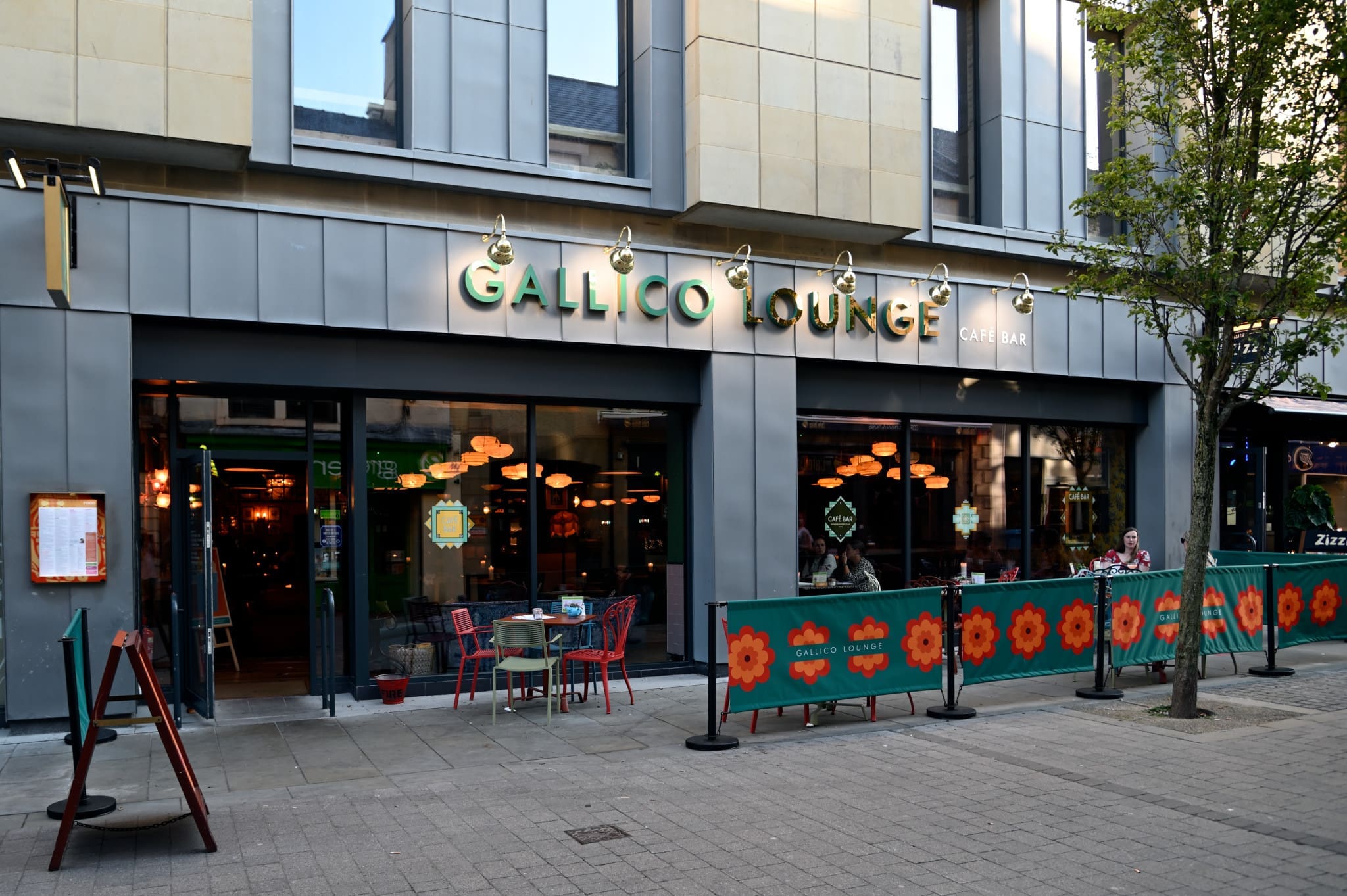 Street view of Gallico Lounge café bar with outdoor seating and flower-patterned barriers; large windows reveal warm hanging lights inside. A tree shades the pavement where people relax in front of Gallico’s inviting atmosphere.