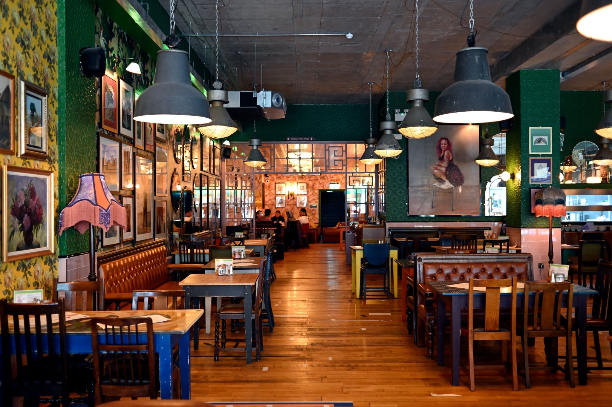 Colorful, eclectic restaurant interior with mismatched chairs, wooden tables, Gallico-inspired framed art on the walls, leather seating, and large industrial pendant lights hanging from a concrete ceiling.