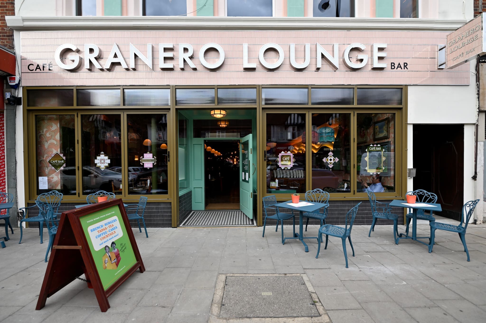 The exterior of Granero Lounge café bar, with large windows, a mint green door, and outdoor seating featuring blue chairs and tables. An A-frame sign is displayed on the pavement near the Granero entrance.