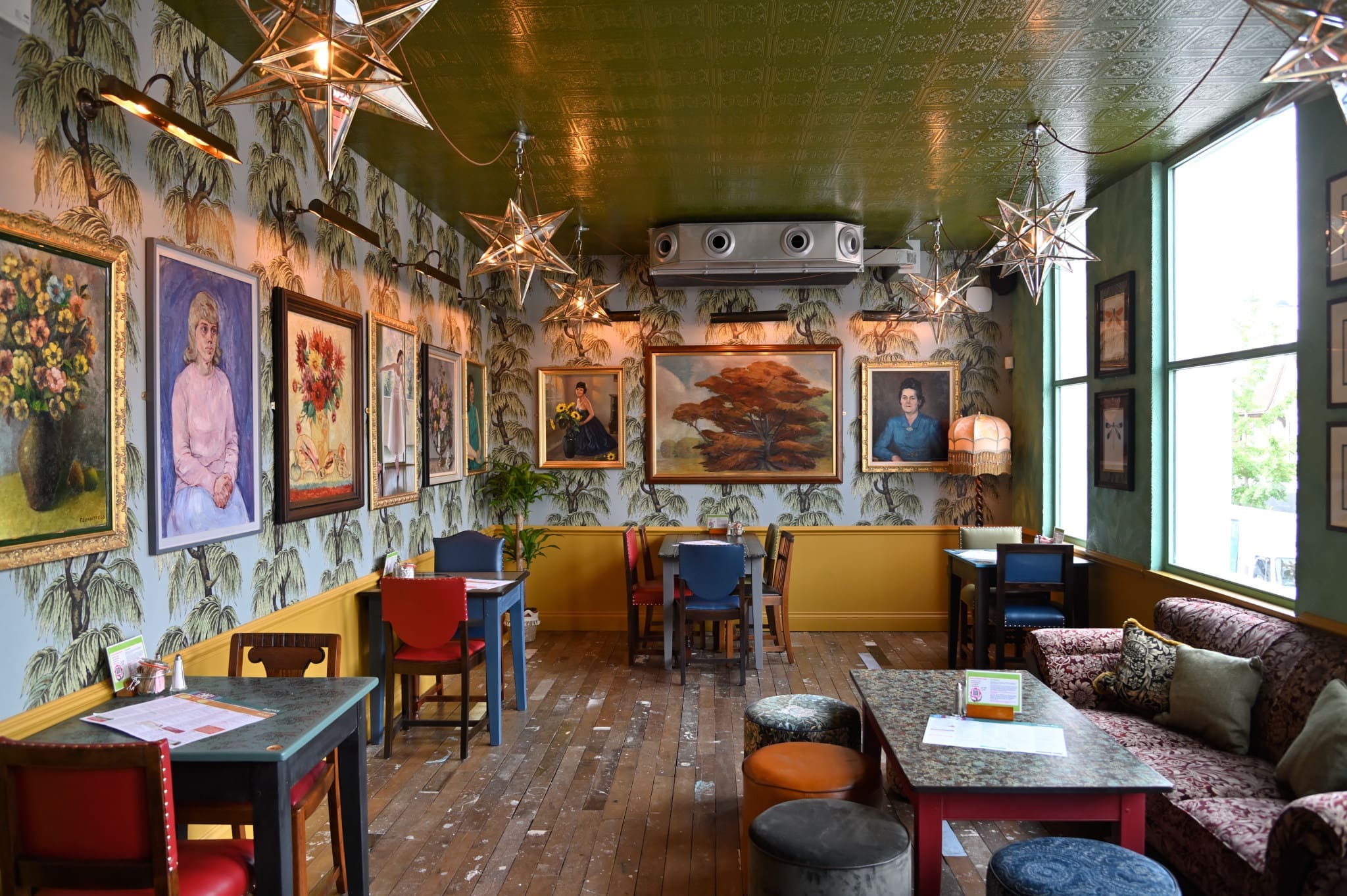 A cozy, eclectic cafe—Granero—features patterned wallpaper, colorful chairs, star-shaped pendant lights, framed artwork, wooden floors, and a large window filling the space with natural light. Tables and couches create a welcoming atmosphere.