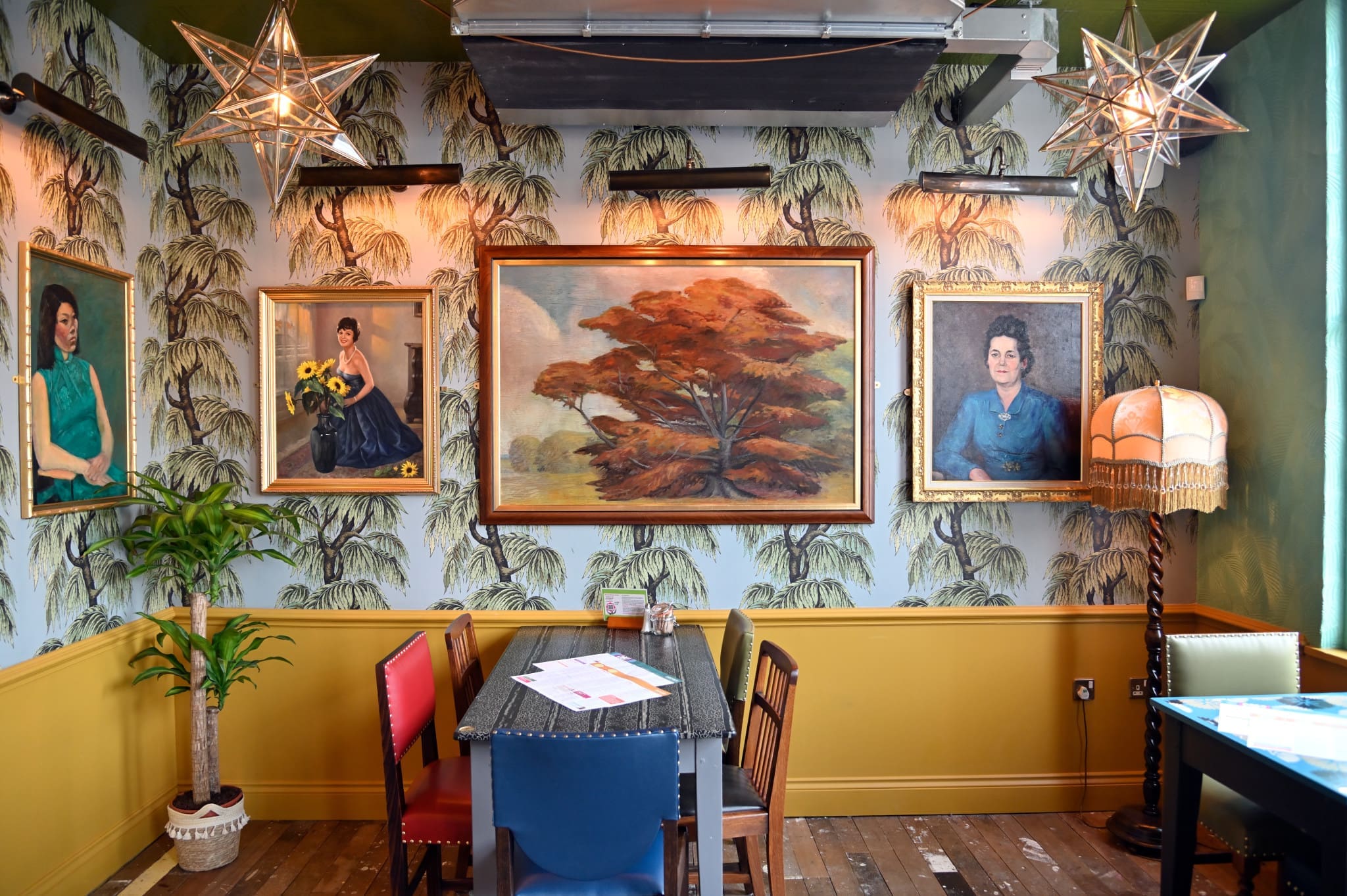 Colorful, eclectic Granero dining area with patterned wallpaper, star-shaped pendant lights, mismatched chairs, vintage portraits, a large tree painting, and a tall lamp in the corner create a cozy, artistic atmosphere.