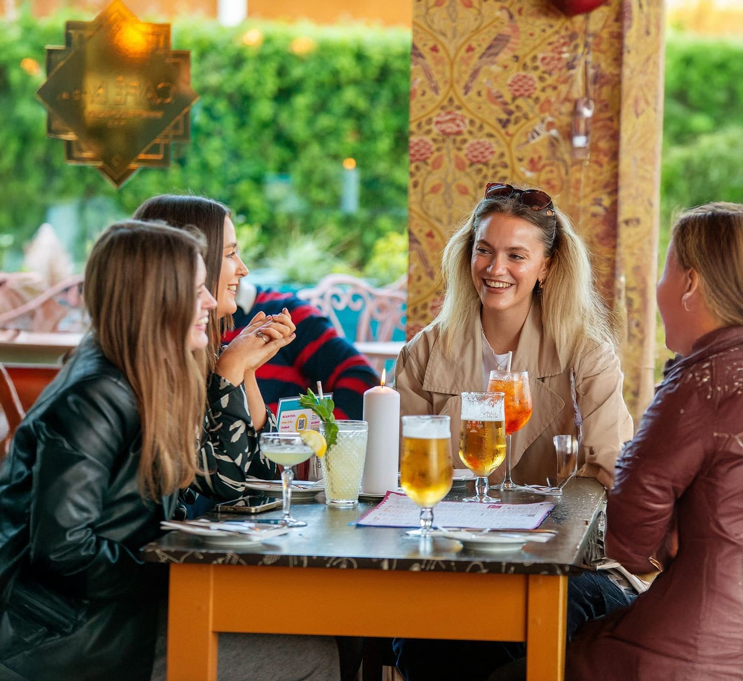 Four women sit around a table outdoors at a lounge, smiling and talking. Drinks, including beer and cocktails, are on the table. The bright, colorful setting features lush greenery in the background.