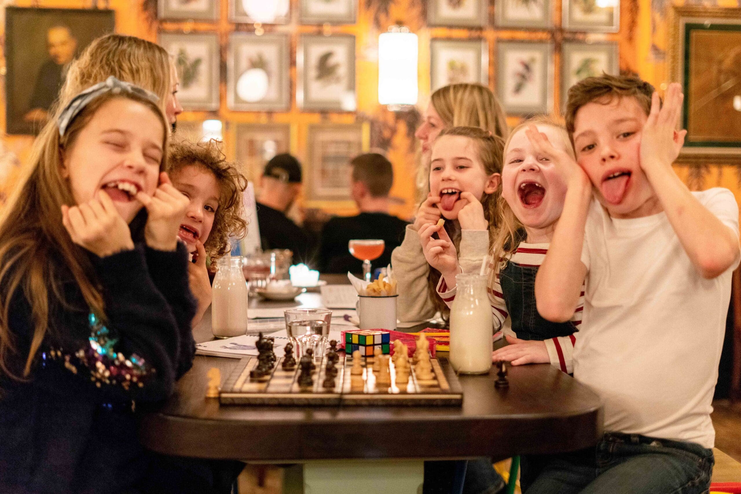 Five children sit around a table making funny faces. There are drinks, a chessboard, and a Rubik’s cube on the table. The background shows framed pictures on a warmly lit wall in a cozy restaurant or cafe.