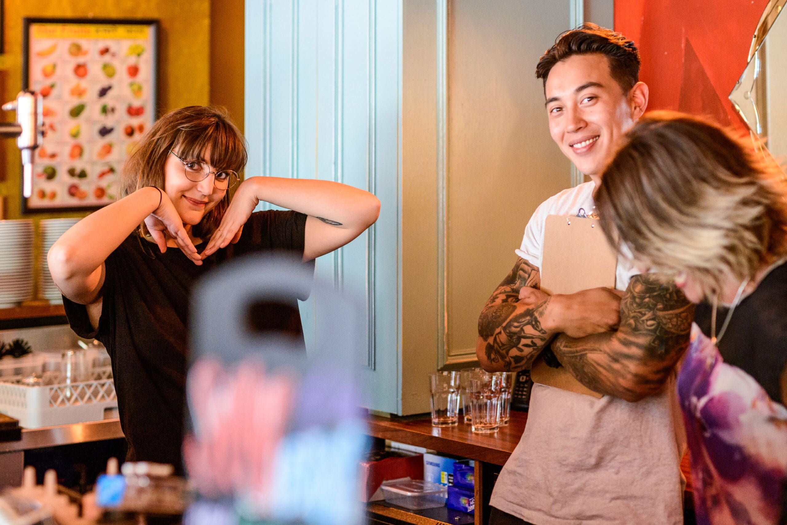 Three people stand behind a counter, smiling. One woman poses with her hands under her chin, another person looks downward, and a man with tattoos holds a clipboard, smiling at the camera. Glasses and dishes are in the background.