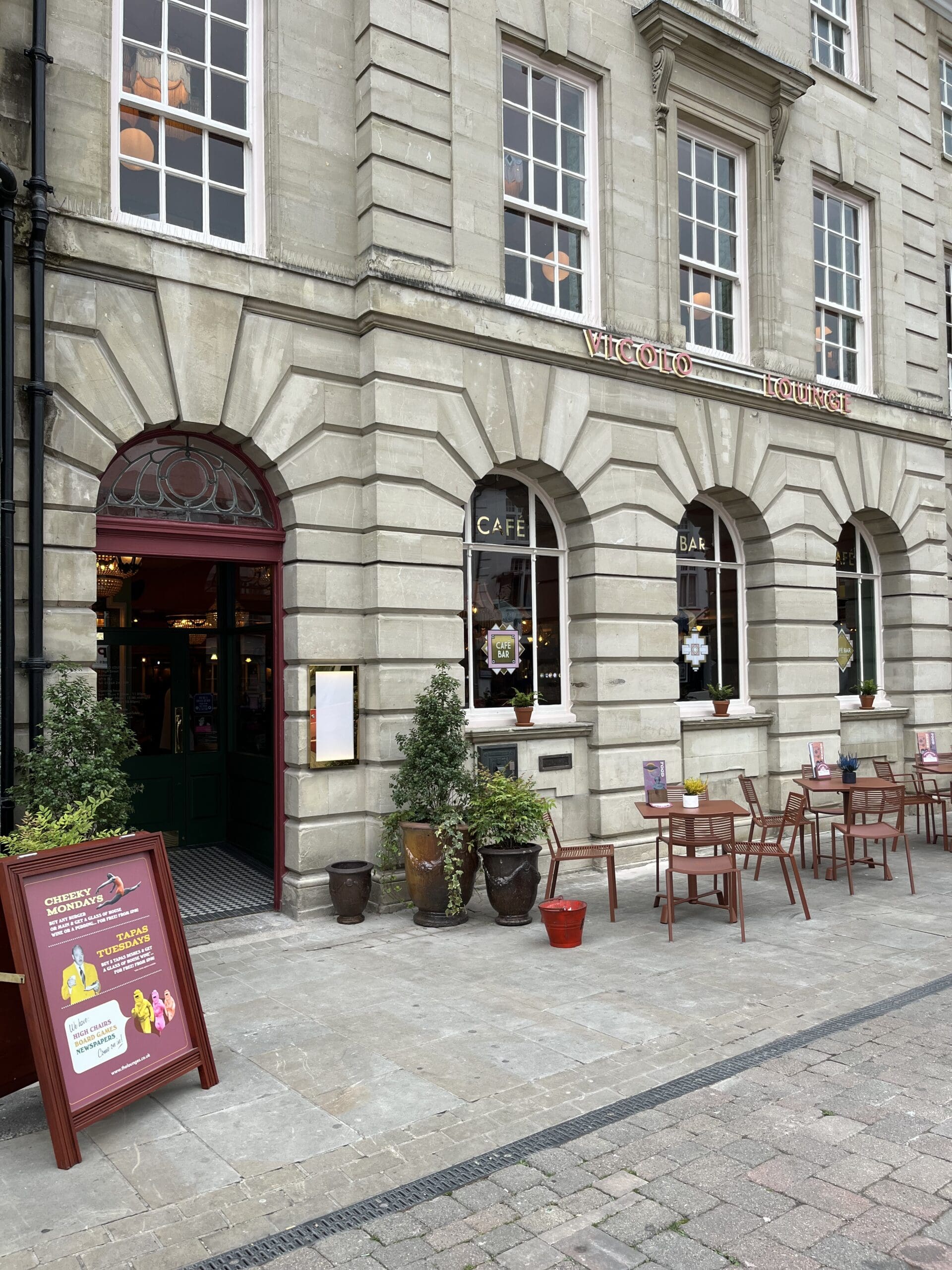 A stone building with arched windows houses Marinero Café and Bar. Potted plants and outdoor tables with chairs line the pavement, while signs with information and posters stand near the entrance.