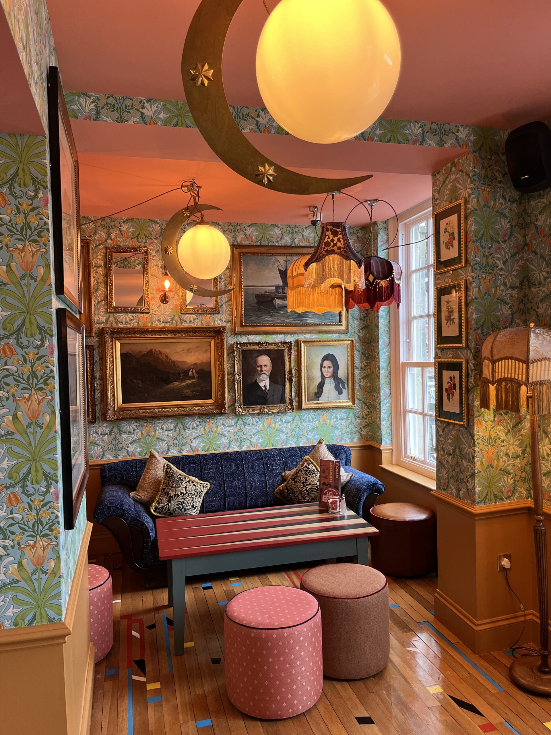 A cozy, colorful room with patterned wallpaper, vintage portraits, and ornate lamps. A blue marinero-style sofa with patterned cushions sits against the wall, surrounded by round pink stools and a striped coffee table. Warm lighting creates an inviting atmosphere.