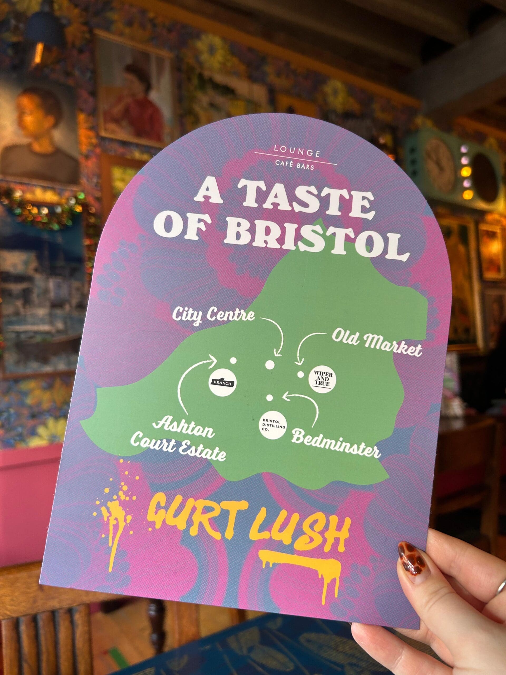 A hand holds a colorful menu reading "A Taste of Bristol" with locations: City Centre, Old Market, Ashton Court Estate, and Bedminster, against a vibrant, decorative cafe background. The phrase "Gurt Lush" is at the bottom.