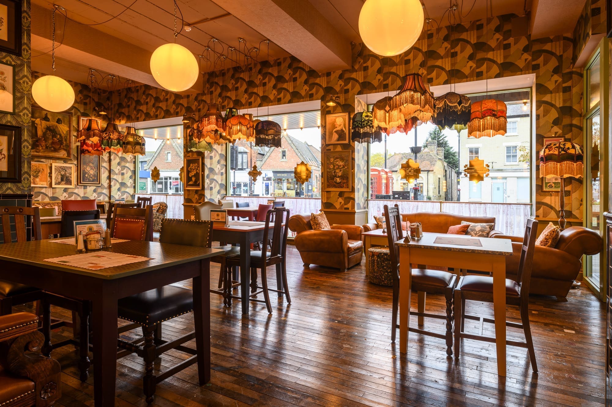 Cozy, vintage-style café interior with patterned wallpaper, wooden floors, eclectic Ivo lampshades, and large windows letting in natural light. Various tables and chairs are arranged for seating.