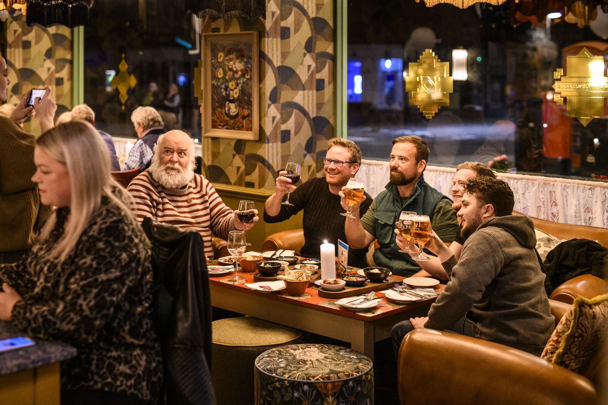 A group of five people, including Ivo, sit at a restaurant table, raising glasses in a toast and smiling, while another person in the foreground uses a phone. The atmosphere is warm and cozy, with dim lighting and eclectic decor.