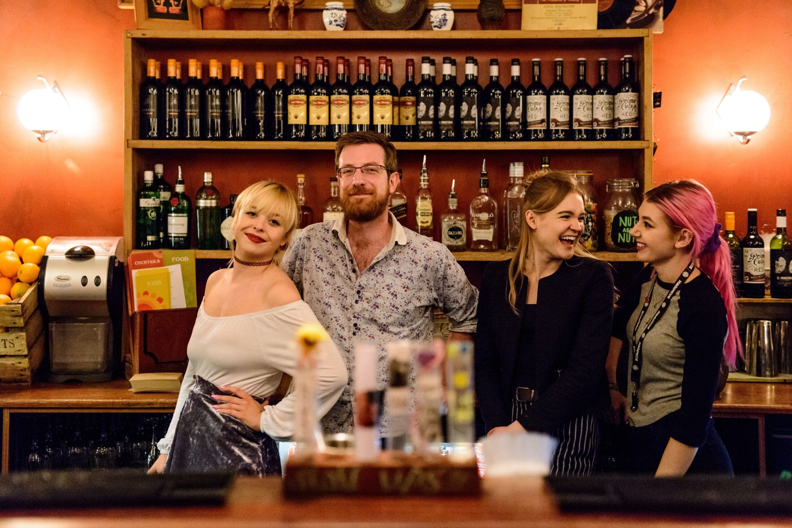 Four people stand behind a bar, smiling and posing. Bottles of wine and spirits are arranged on shelves behind them, hinting at rewarding careers in hospitality. The warm lighting creates a cozy, inviting atmosphere.