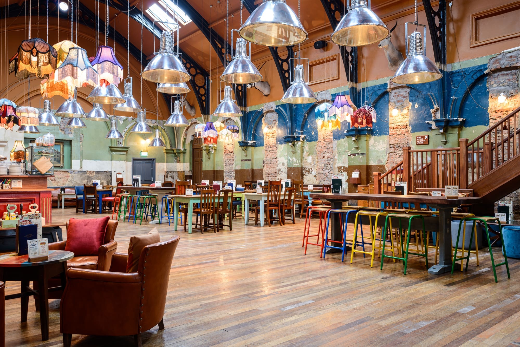 A spacious, eclectic cafe with exposed brick columns, colorful mismatched chairs, wooden tables, and industrial lights—Kino vibes abound under a high ceiling with skylights and various hanging lampshades.